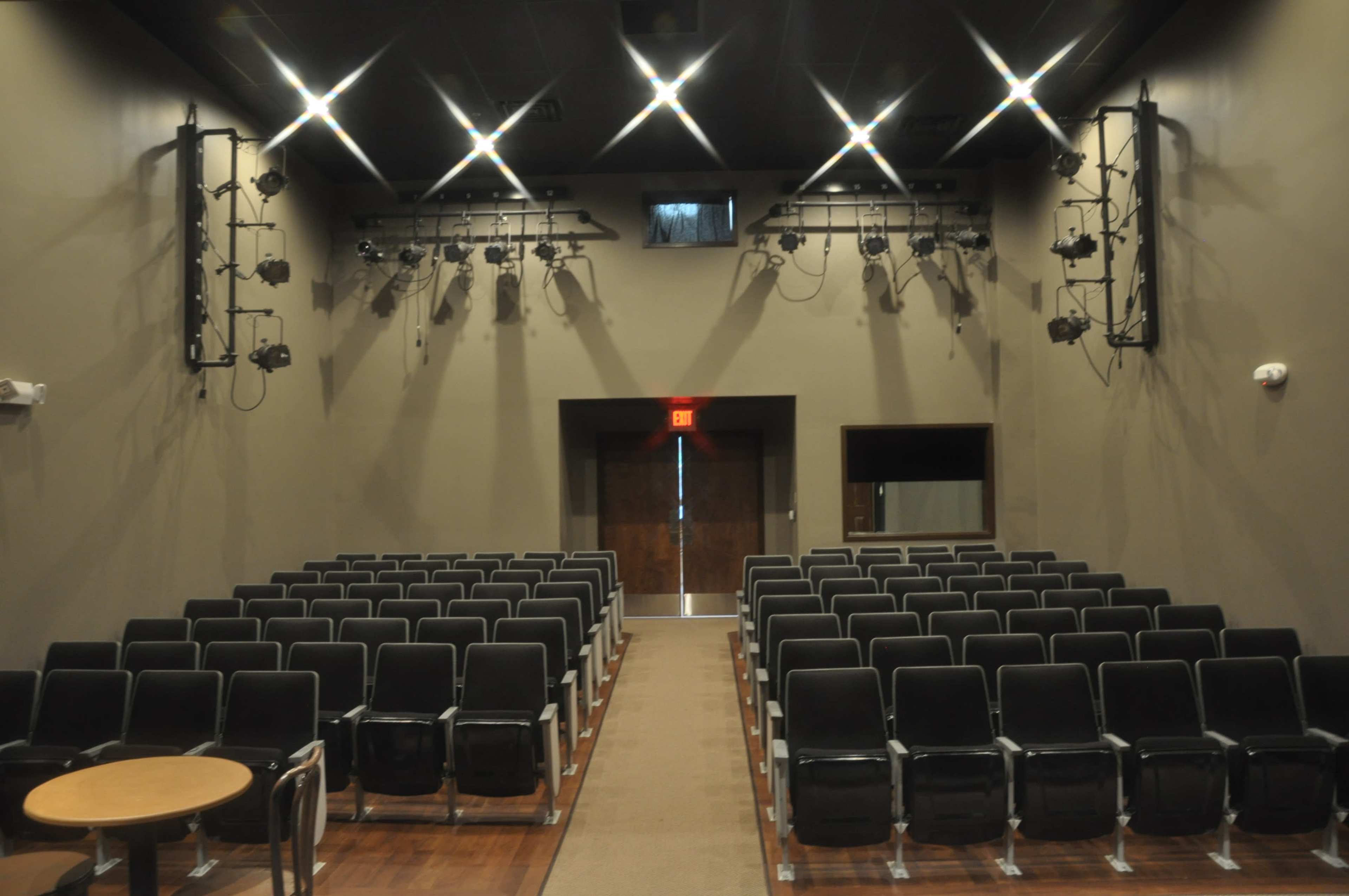 A half-empty theater with rows of black chairs and stage lighting fixtures mounted on the walls.