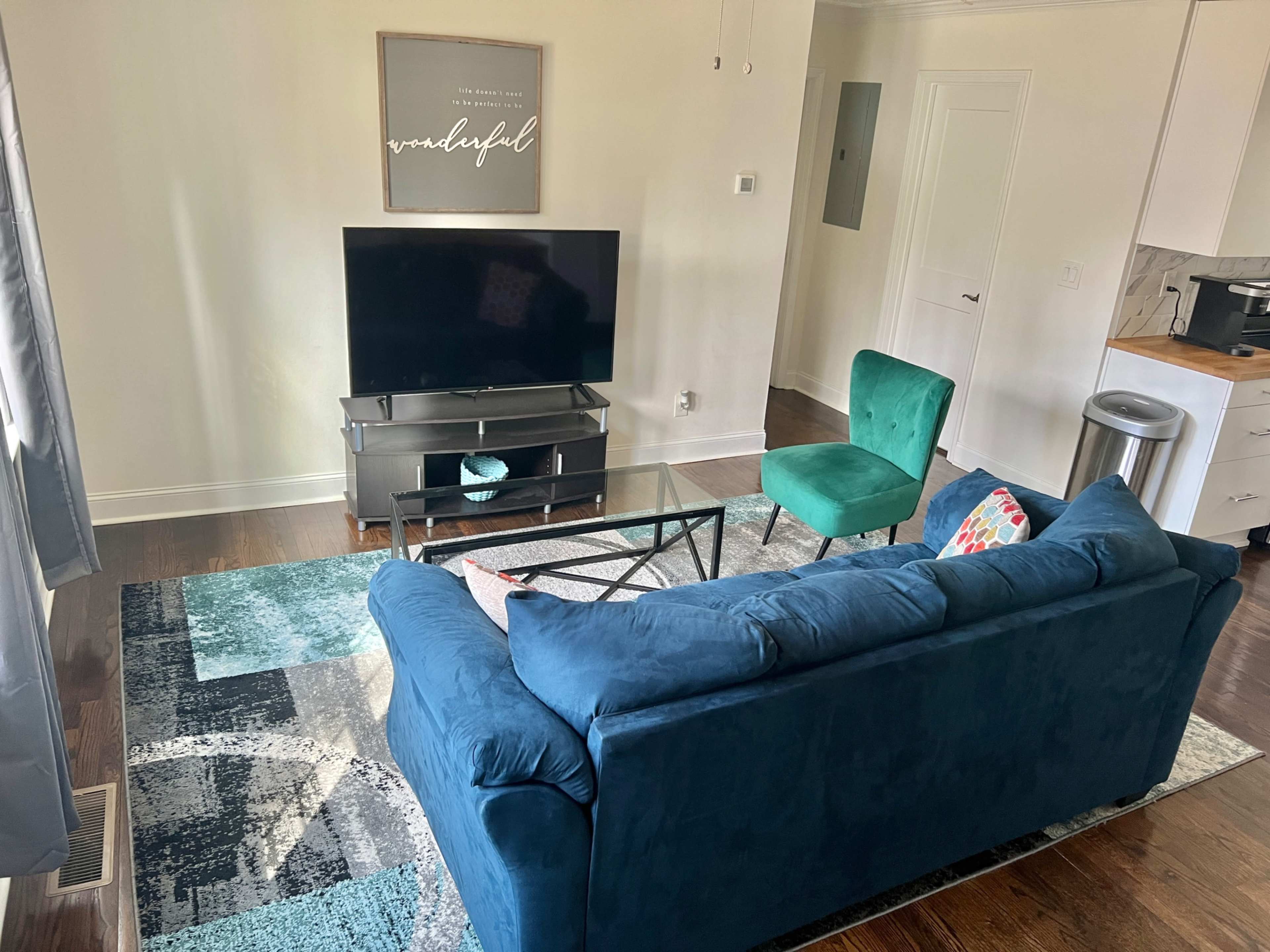A living room features a blue sofa, a green chair, a television on a stand, and a patterned rug.