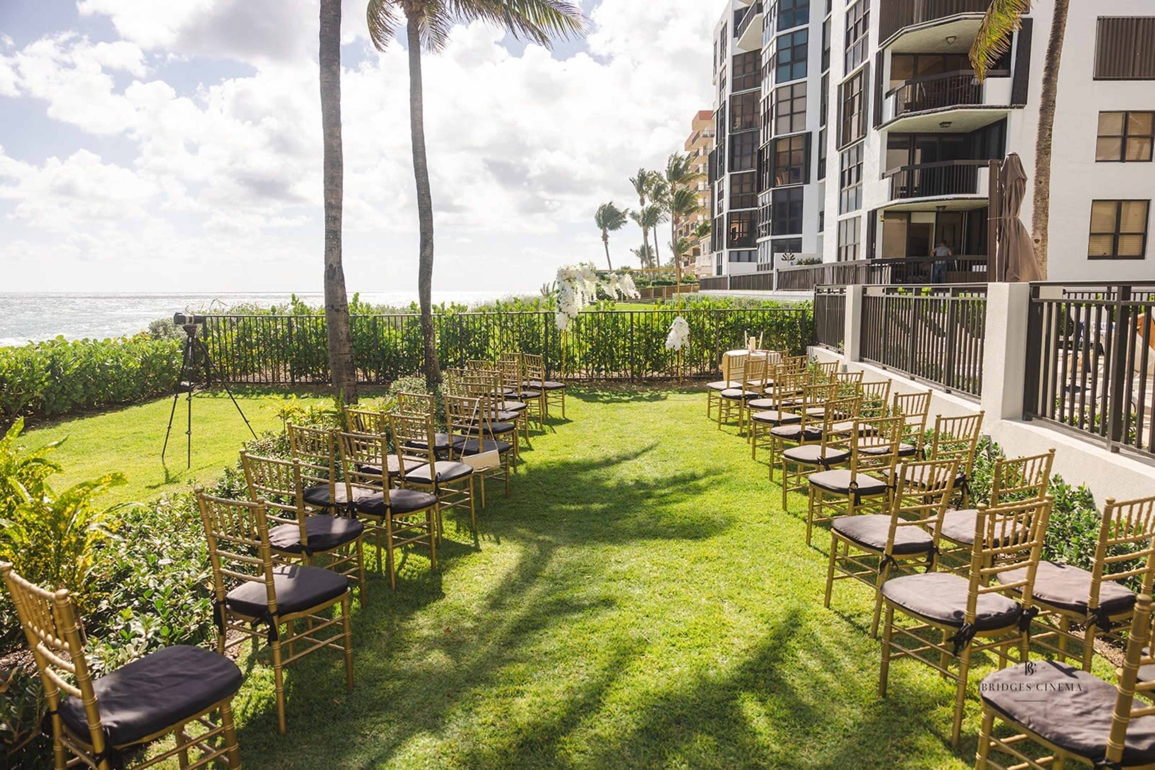 A setting for an outdoor event features rows of gold chairs arranged on grass near a seaside building, with palm trees in the background.