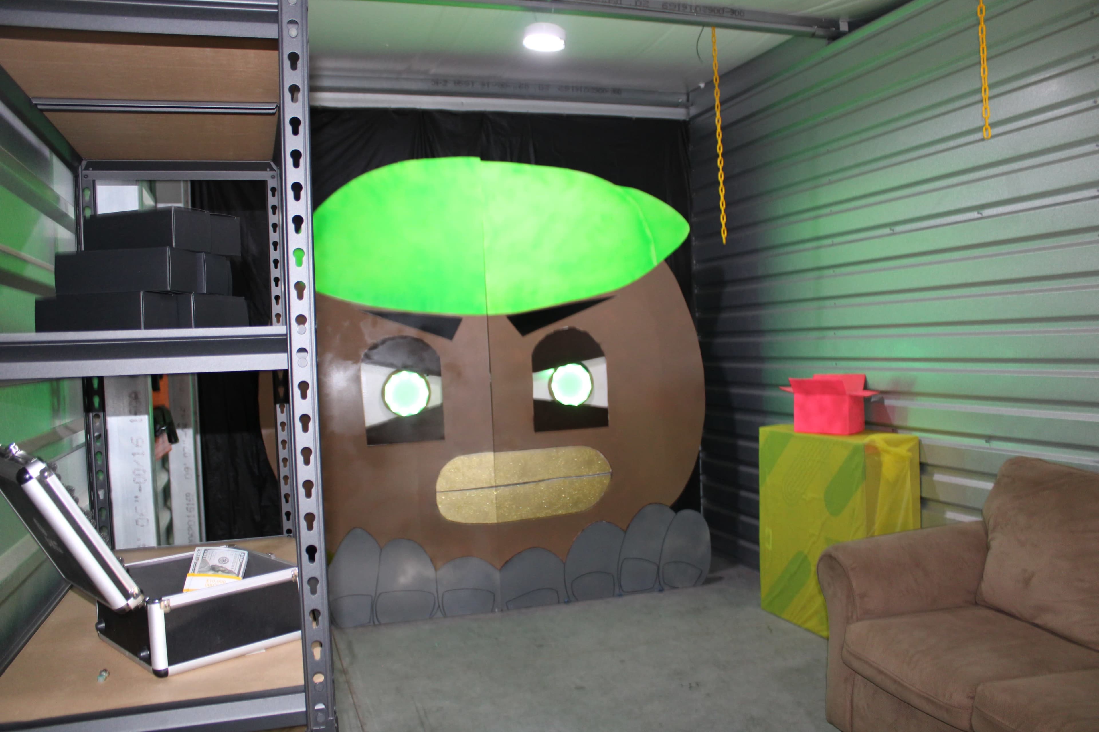 The image shows a storage unit with a large, cartoonish character mural on the wall, a brown and green design, accompanied by a brown couch and colorful boxes in the corner.