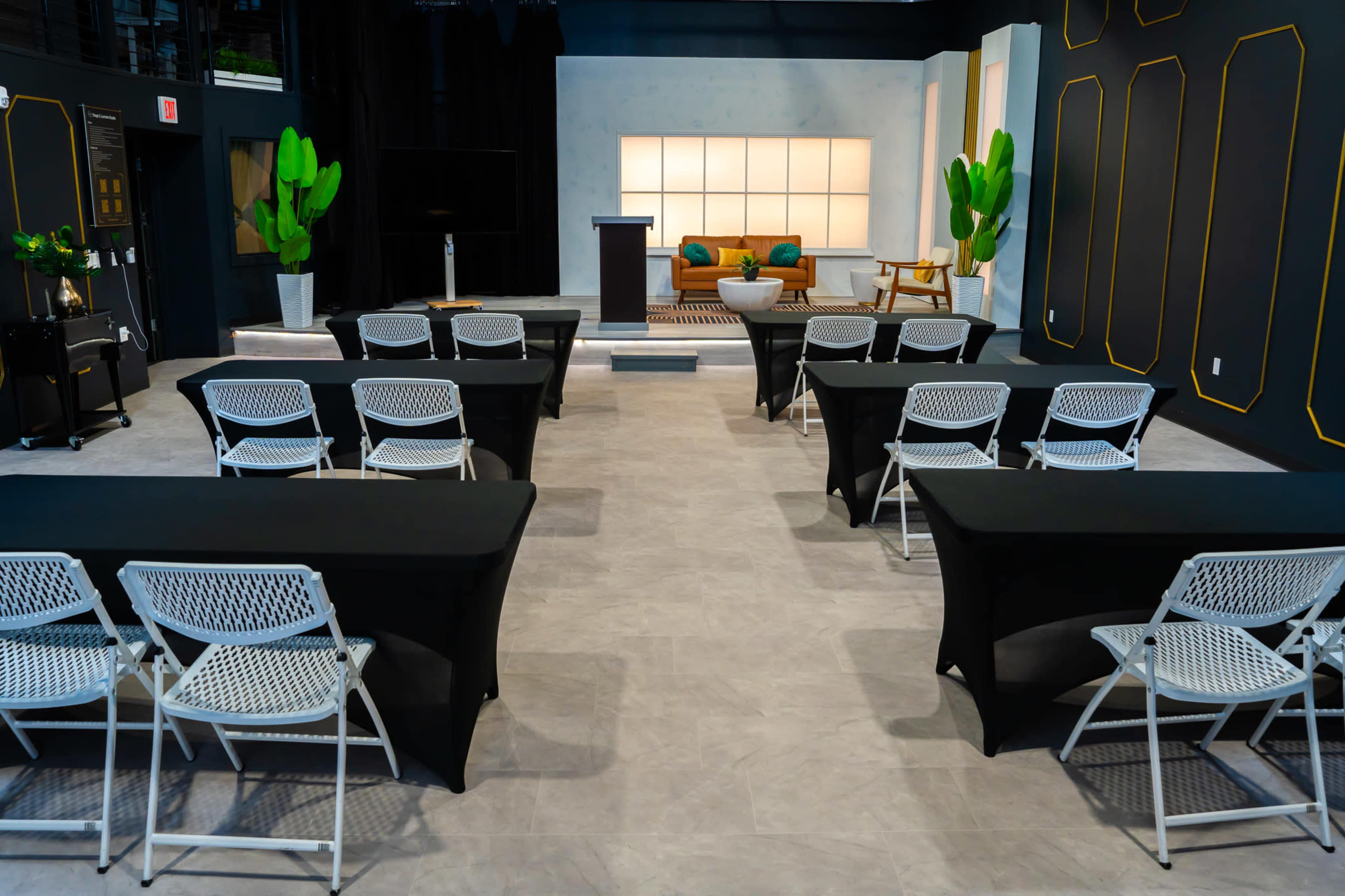 The image shows a conference room setup with rows of white folding chairs and black tables, featuring a podium and a seating area with a couch and plants in the background.