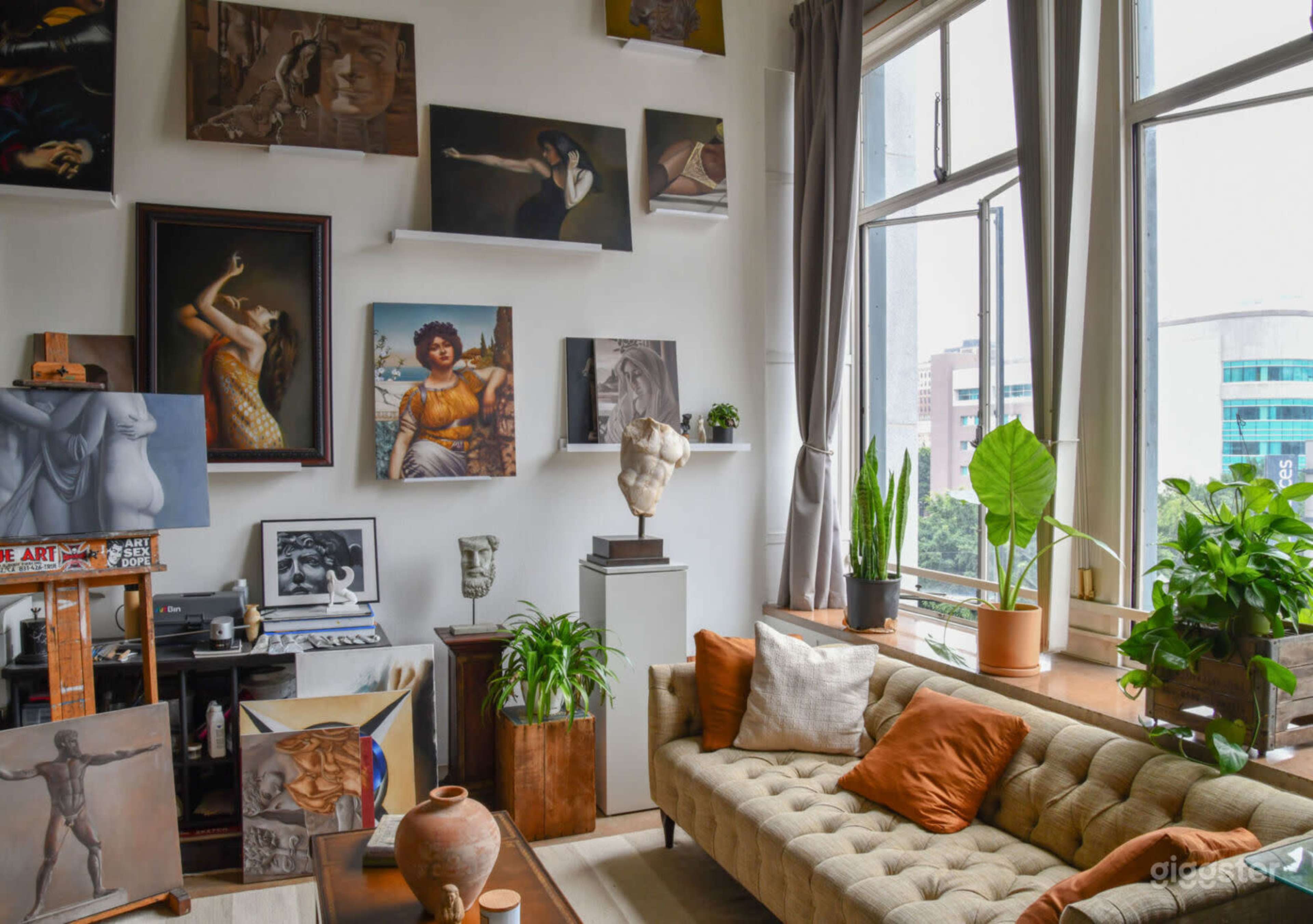 The image shows a cozy art-filled room featuring a beige sofa with orange cushions, various framed artworks on the walls, and plants near a window.