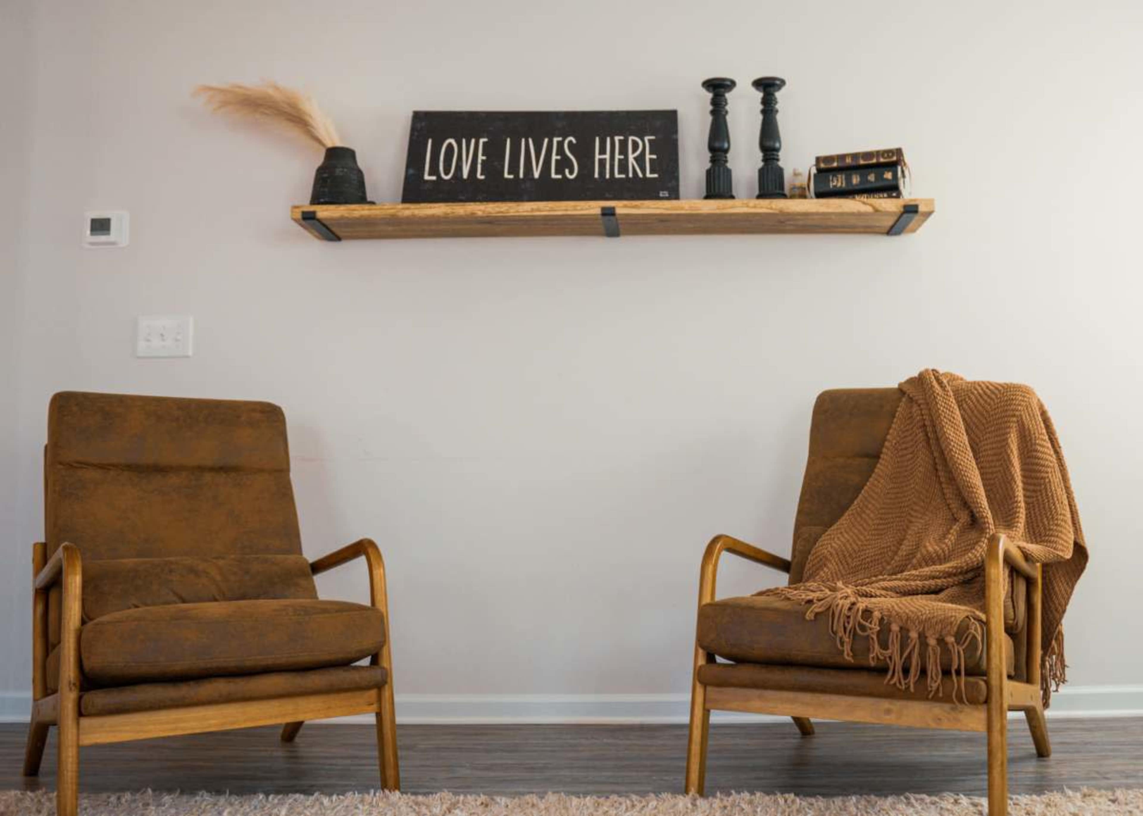 Two mid-century modern chairs with a brown finish are placed facing each other on a rug beneath a wooden shelf holding decorative items and a sign that reads "LOVE LIVES HERE."