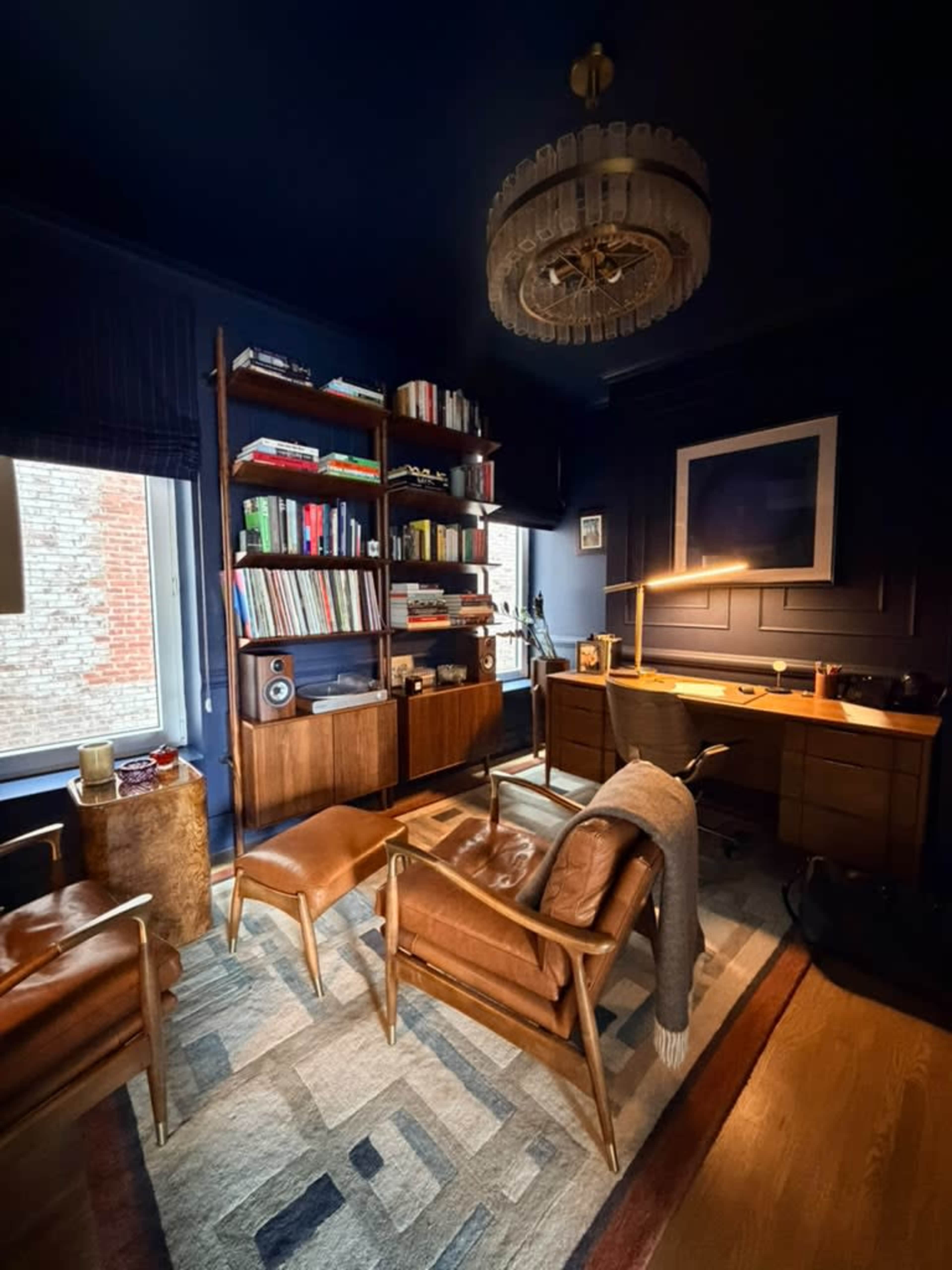 The image shows a styled home office with dark blue walls, wooden furniture, a bookshelf filled with books, and two brown chairs.