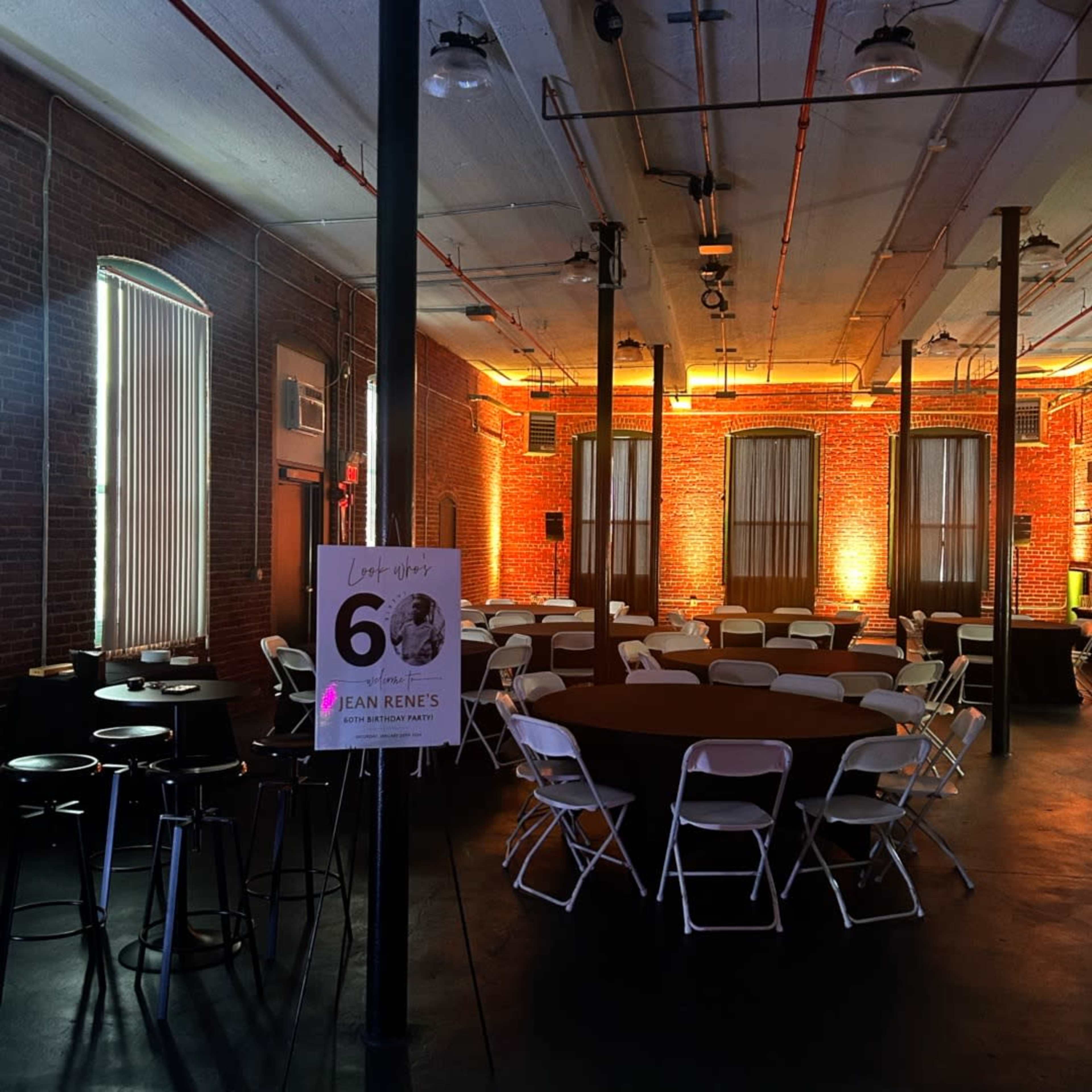 An event space featuring round tables and chairs arranged in a well-lit brick room with large windows and exposed pipes.