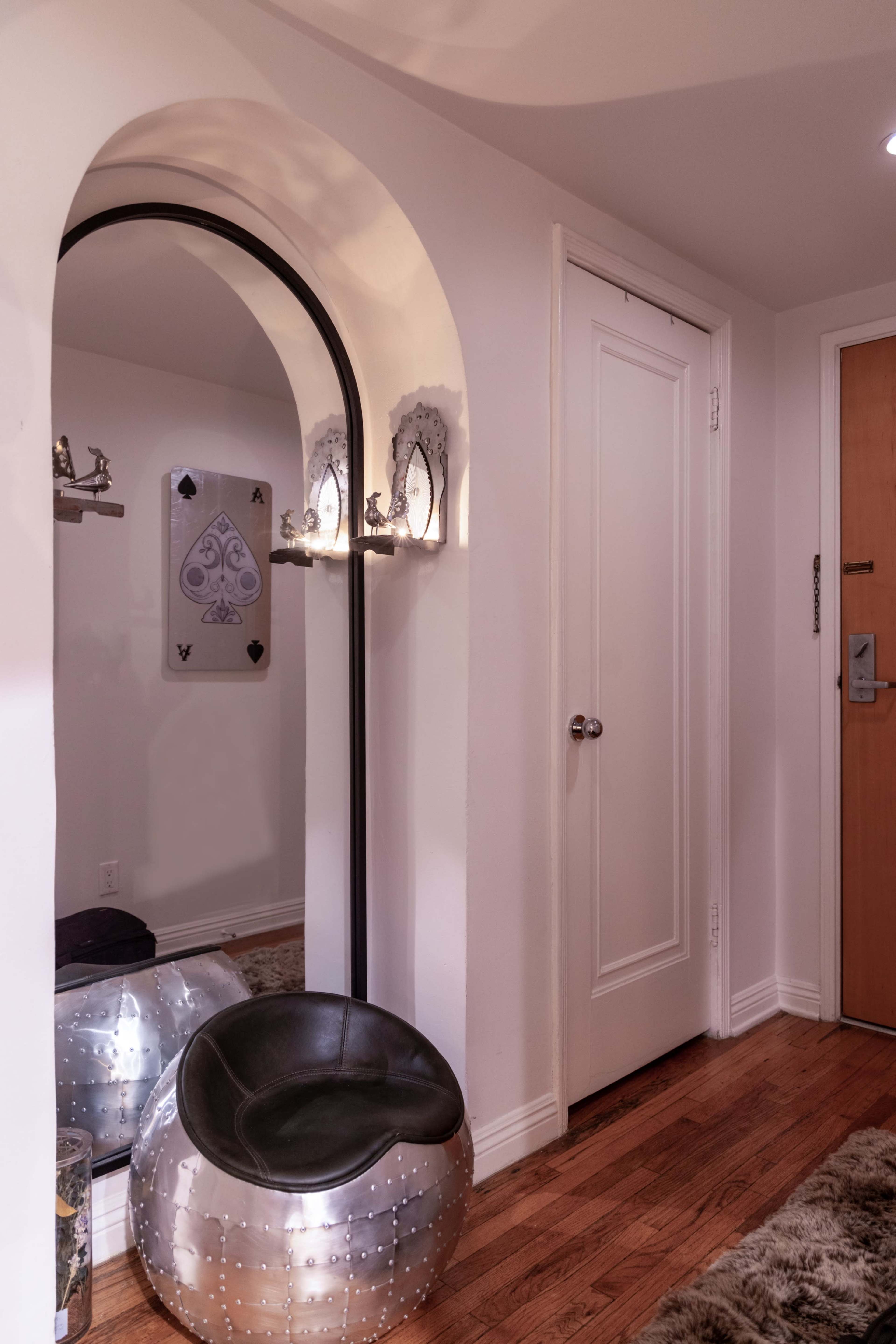 A hallway features a large mirror with an arched top, a sleek silver ottoman, and a closed white door.