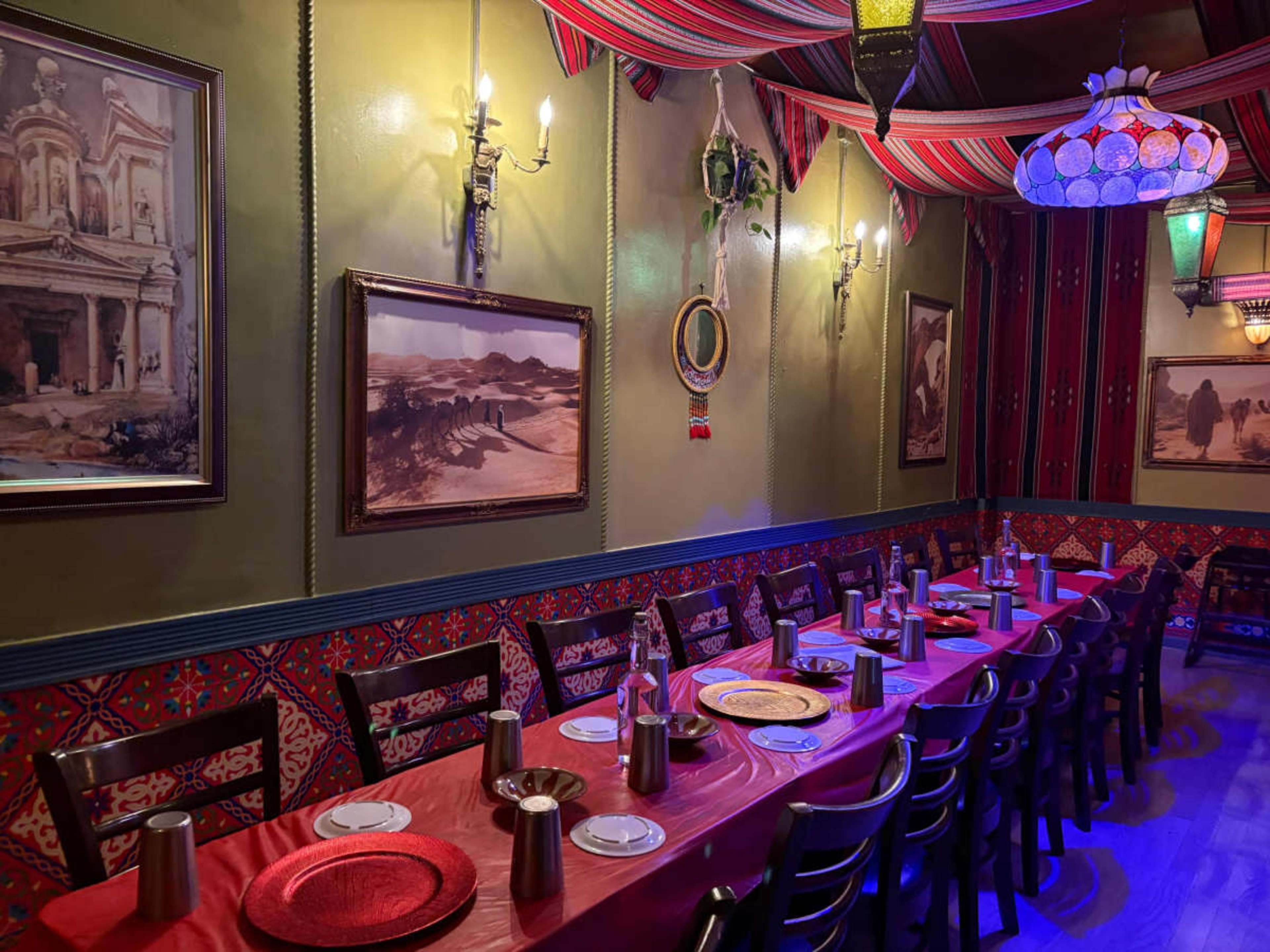 The image shows a dining area of a restaurant featuring a long table set with plates and metal mugs, surrounded by colorful decor and artwork on the walls.