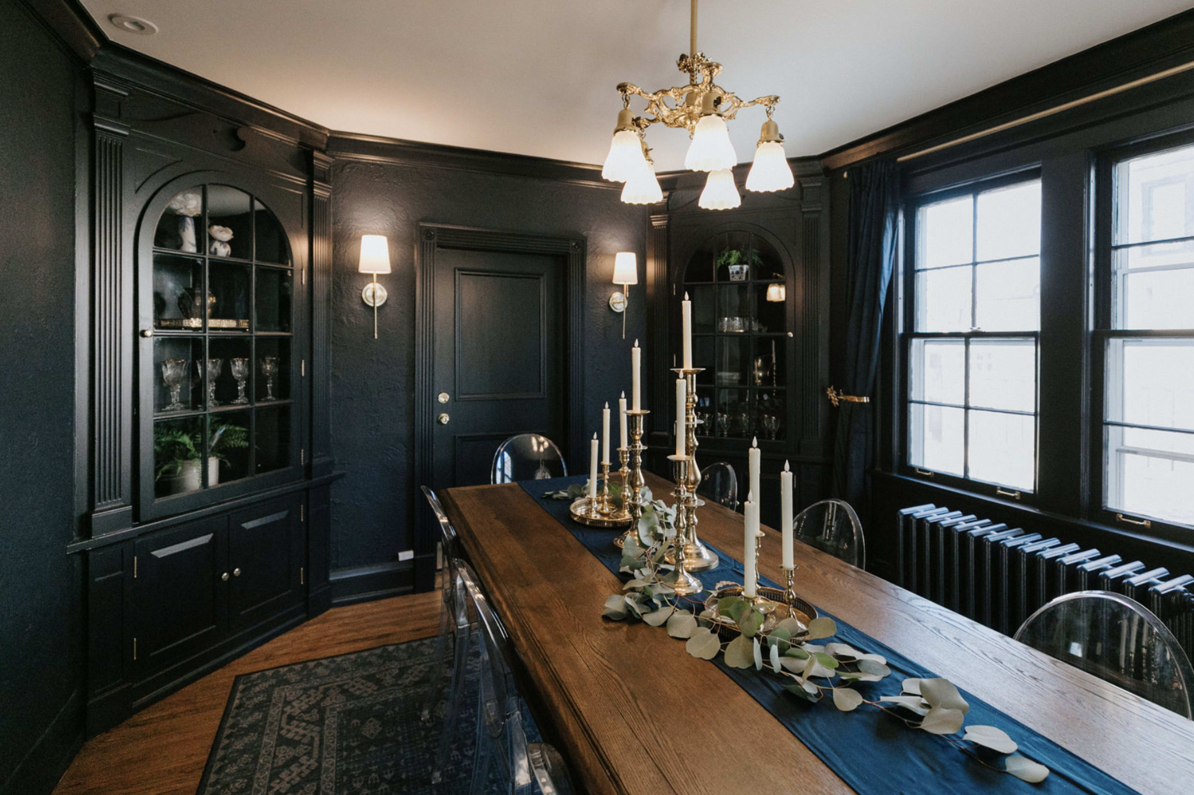 The image shows a dark-paneled dining room featuring a long wooden table with decorative candlesticks and a runner, surrounded by clear acrylic chairs.