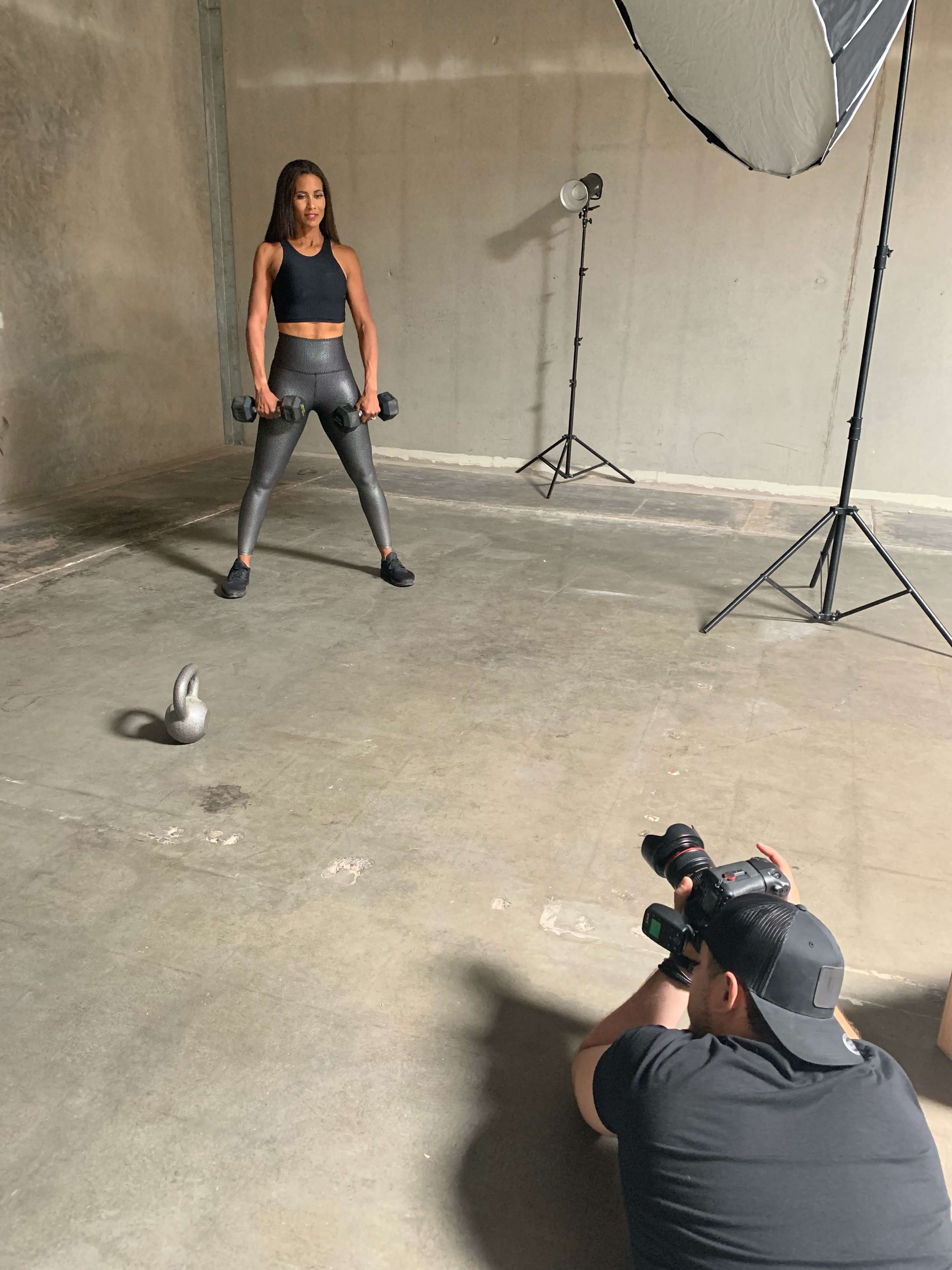 A woman holds dumbbells while a photographer captures her pose in an industrial-style studio.