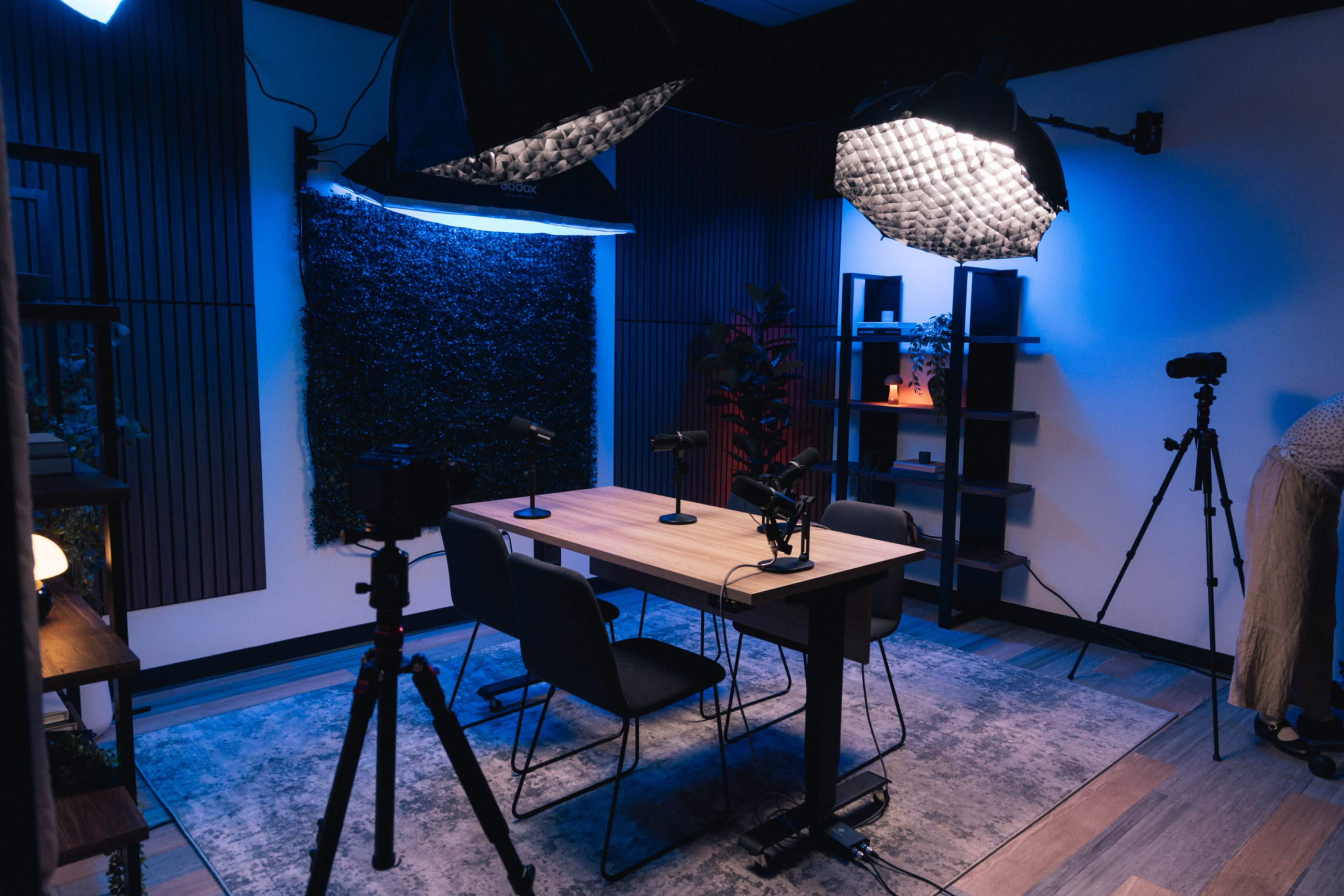 The image shows a recording studio set up with a table, chairs, microphones, and lighting equipment against a backdrop of dark textured walls.