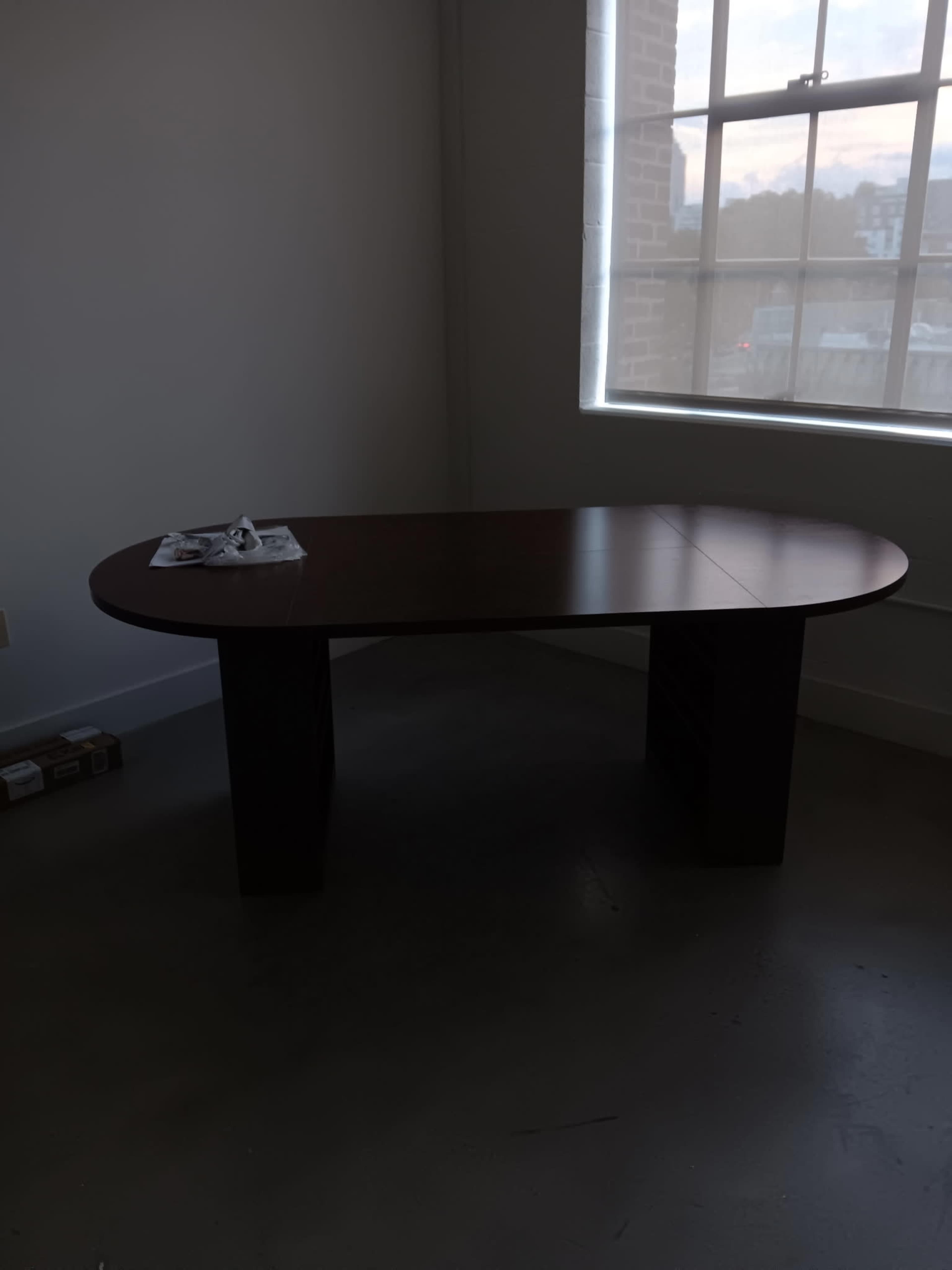 A brown oval table with two rectangular legs is positioned near a large window in a room with light-colored walls and a concrete floor.