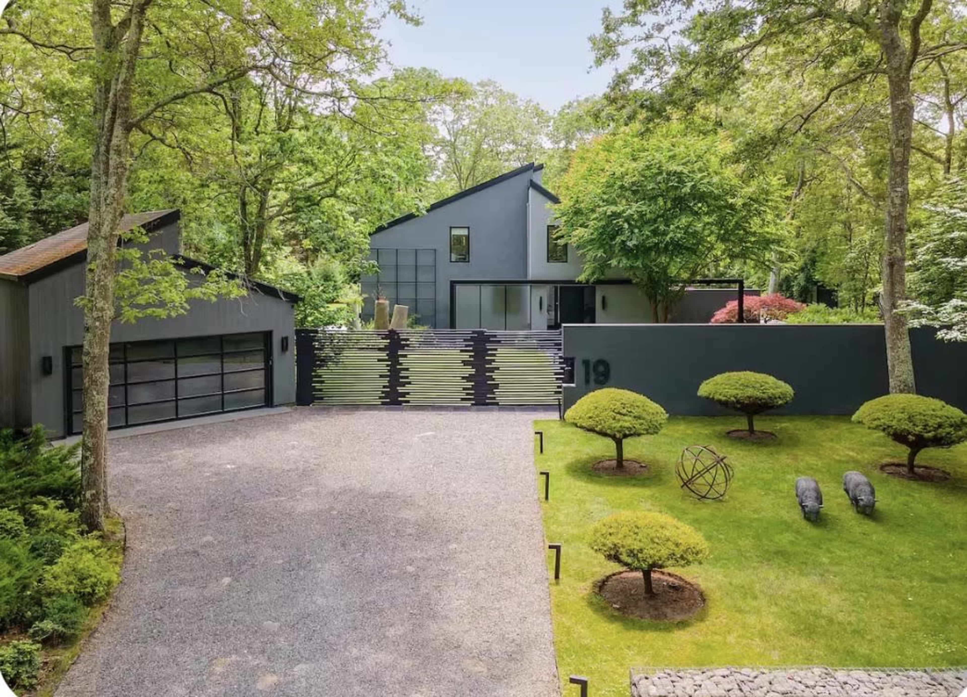 A modern black house surrounded by lush greenery features a gravel driveway, landscaped gardens, and stylized sculptures in the yard.