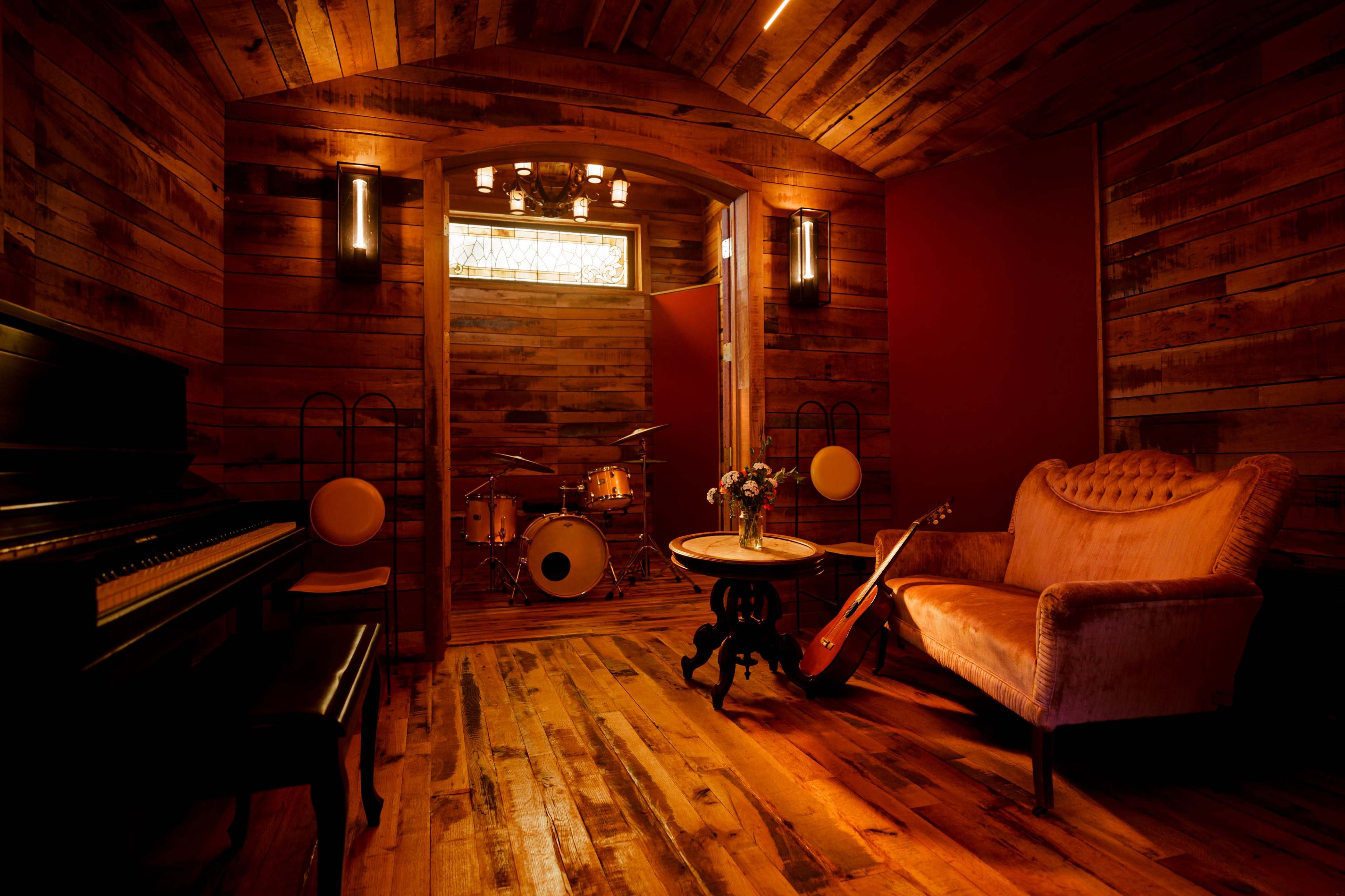 The room features a wooden interior with a piano, a drum set, a vintage-style sofa, and a guitar resting by a small table.
