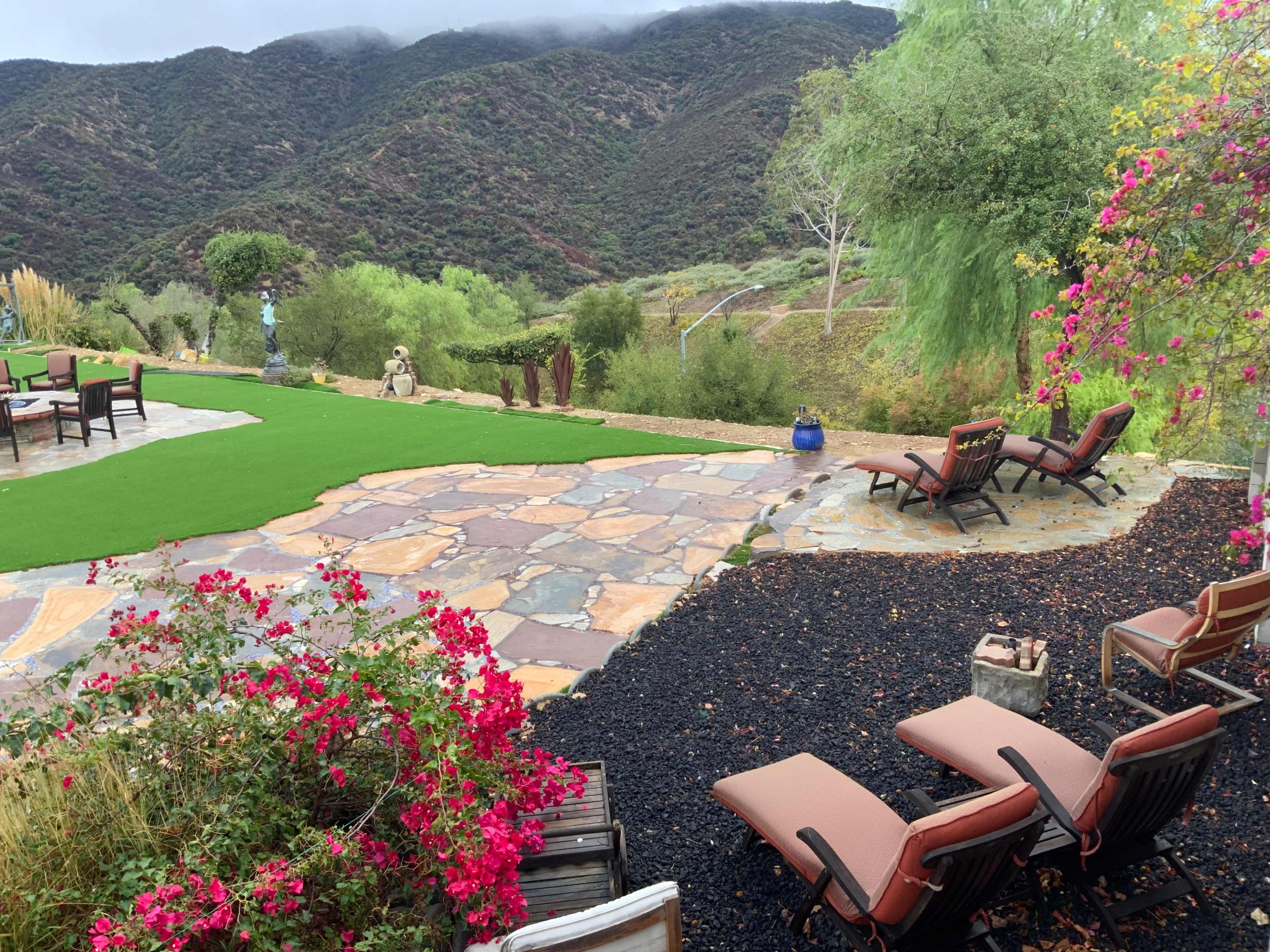 The scene features a patio area with stone pathways, chairs, and vibrant flowers, set against a backdrop of green mountains and cloudy skies.