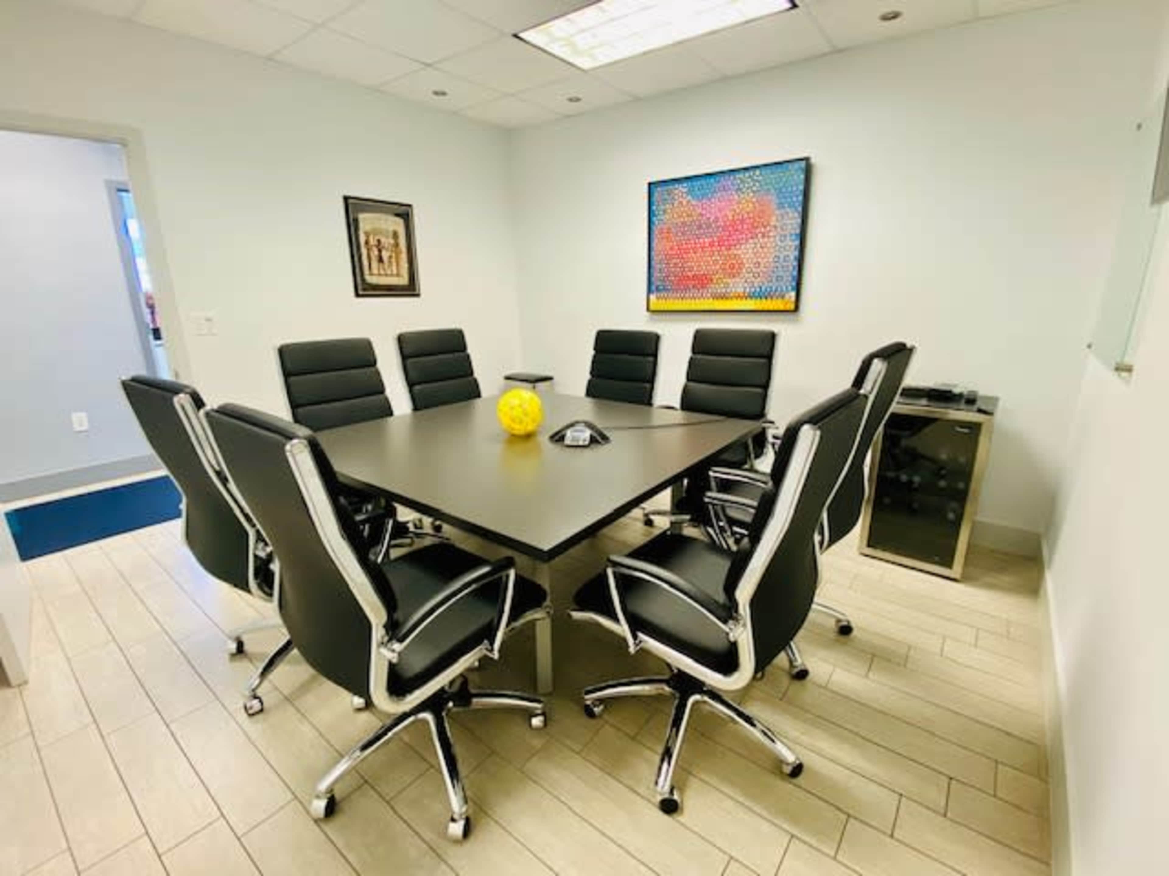 A conference room features a large black table surrounded by eight black office chairs, with a yellow decorative object in the center and colorful artwork on the wall.