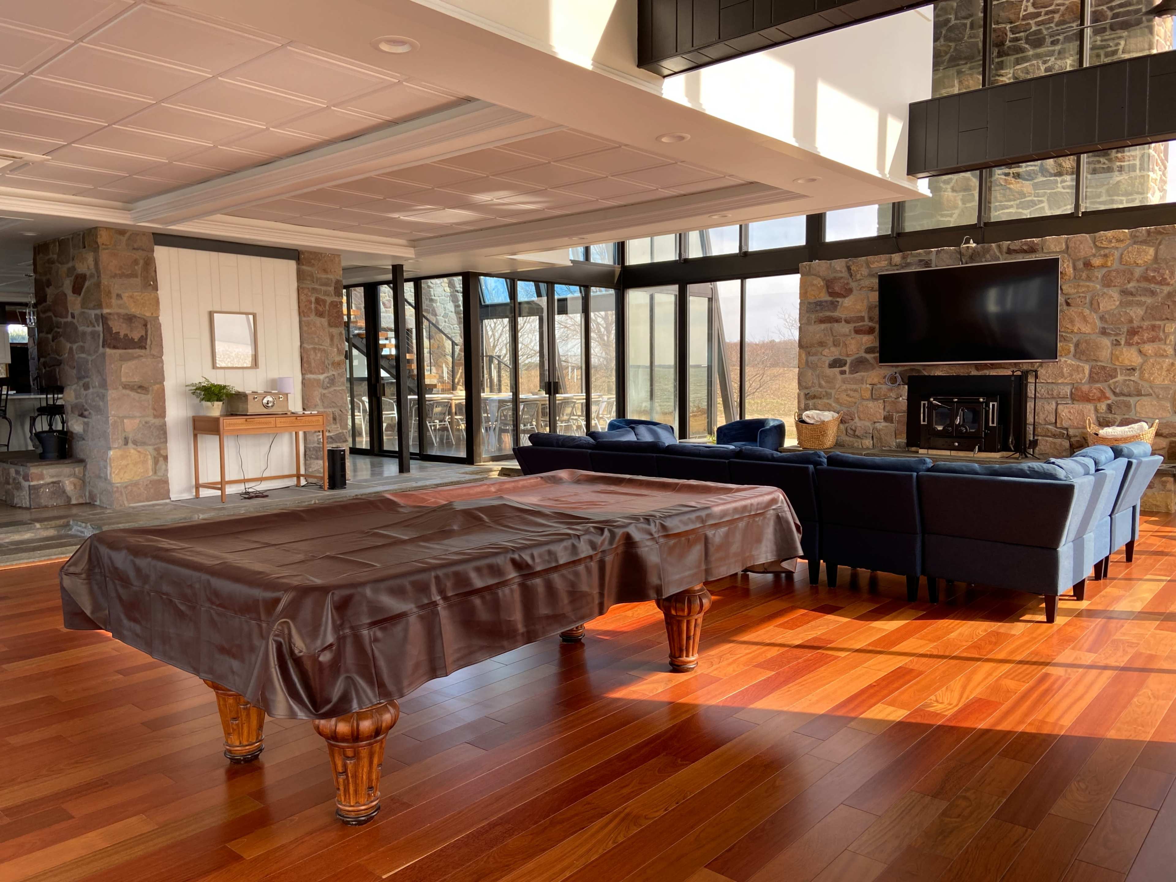 A modern indoor space featuring a pool table covered with a brown cloth, surrounded by large windows, a stone fireplace, and a seating area with blue sofas.