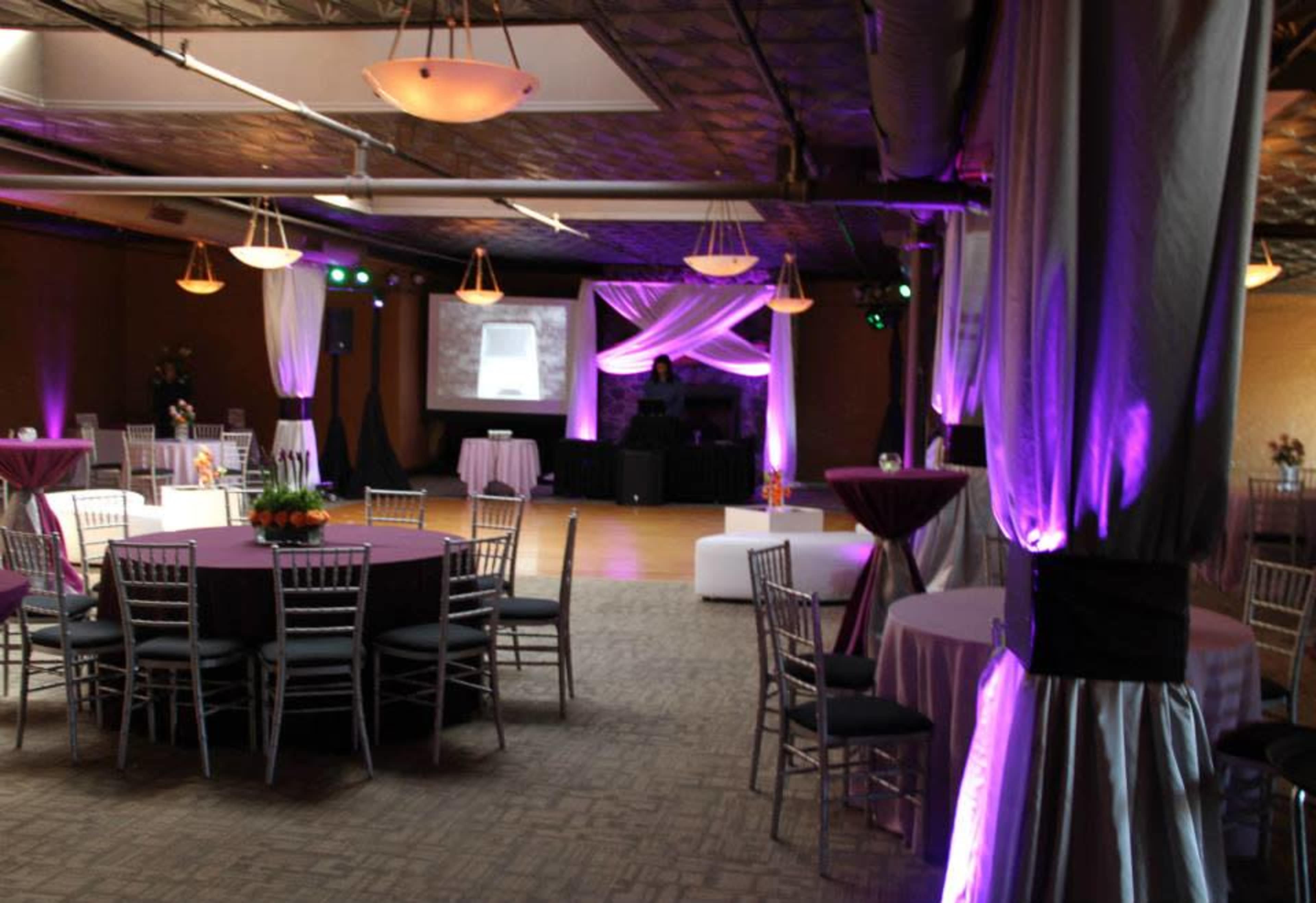 A decorated event space featuring several round tables with chairs, ambient lighting, and a DJ setup in the background.