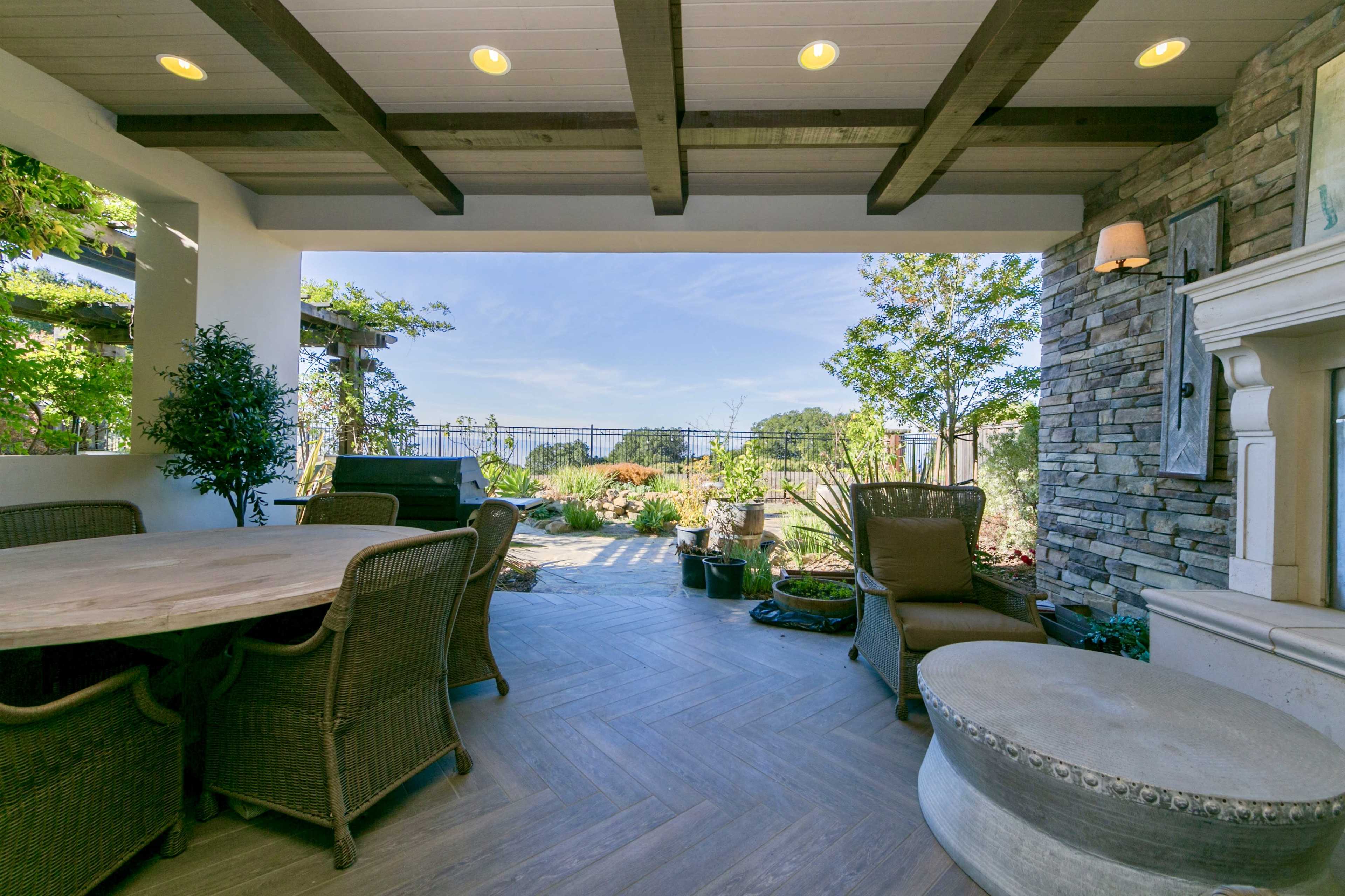 A covered outdoor patio with a wooden table and wicker chairs, overlooking a landscaped garden.