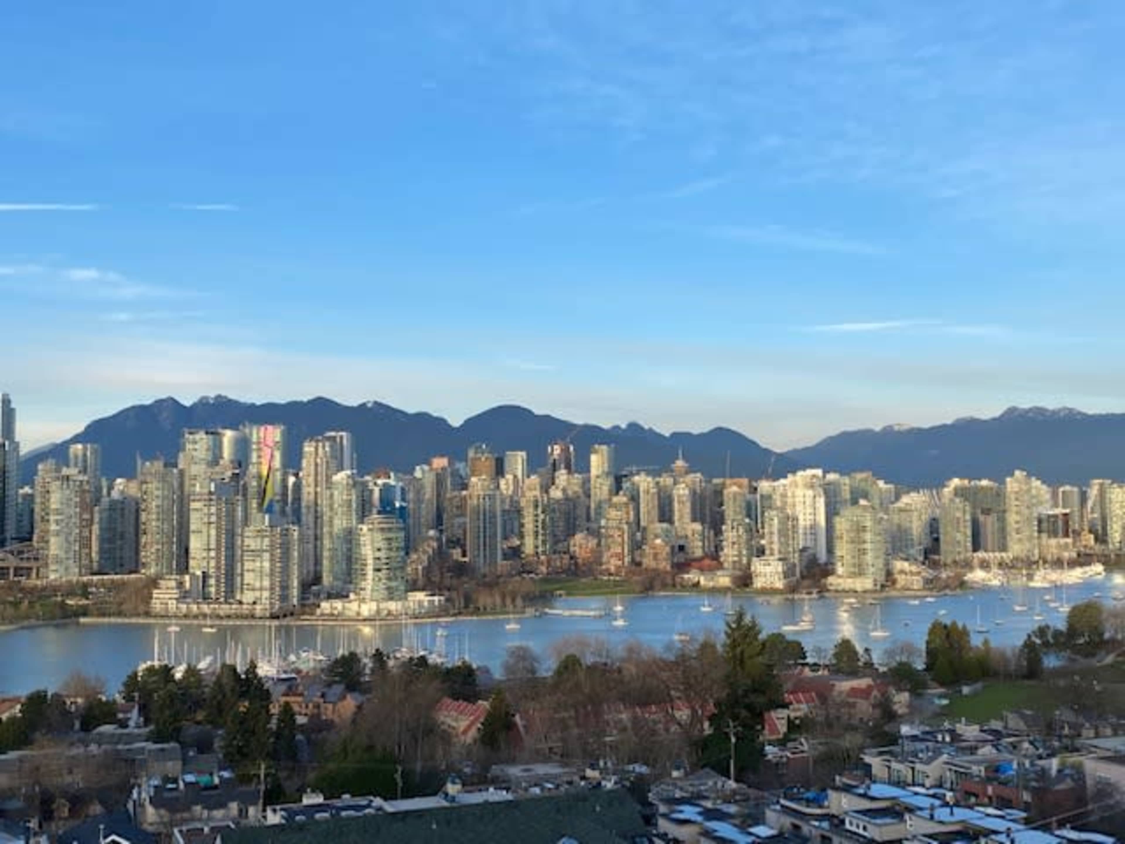 A skyline of tall buildings along a body of water with mountains in the background under a clear blue sky.