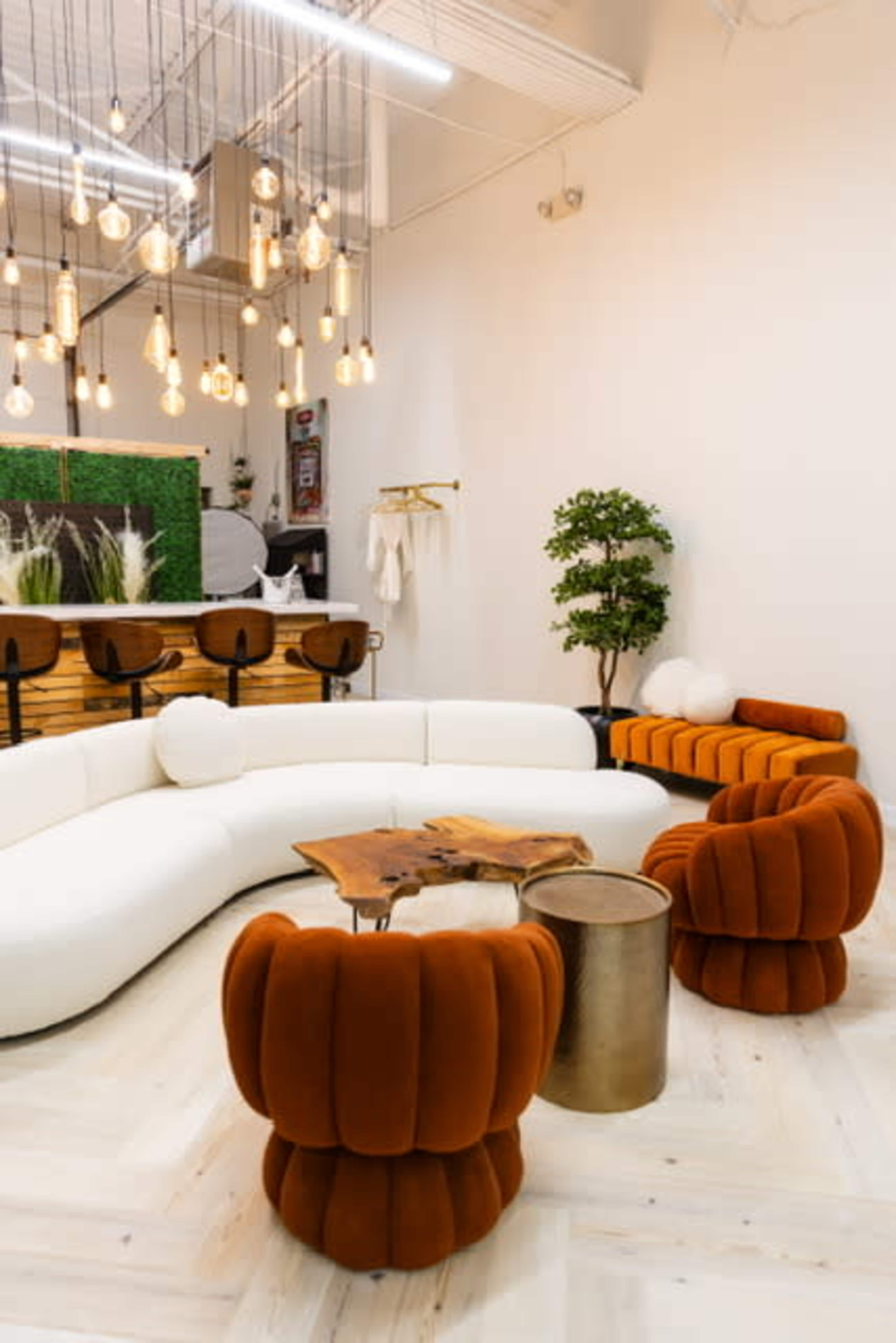 The image shows a modern lounge area featuring a white curved sofa, orange upholstered chairs, a wooden table, and decorative lighting above.