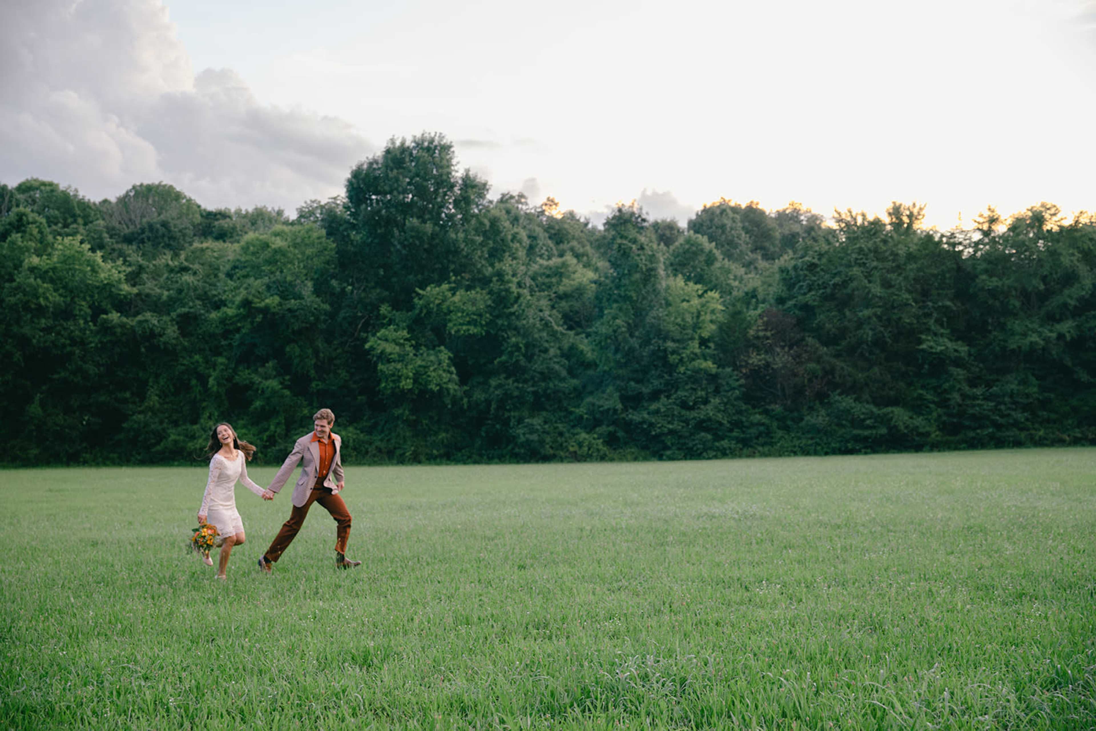 A couple dressed in wedding attire joyfully runs across a grassy field with trees in the background.