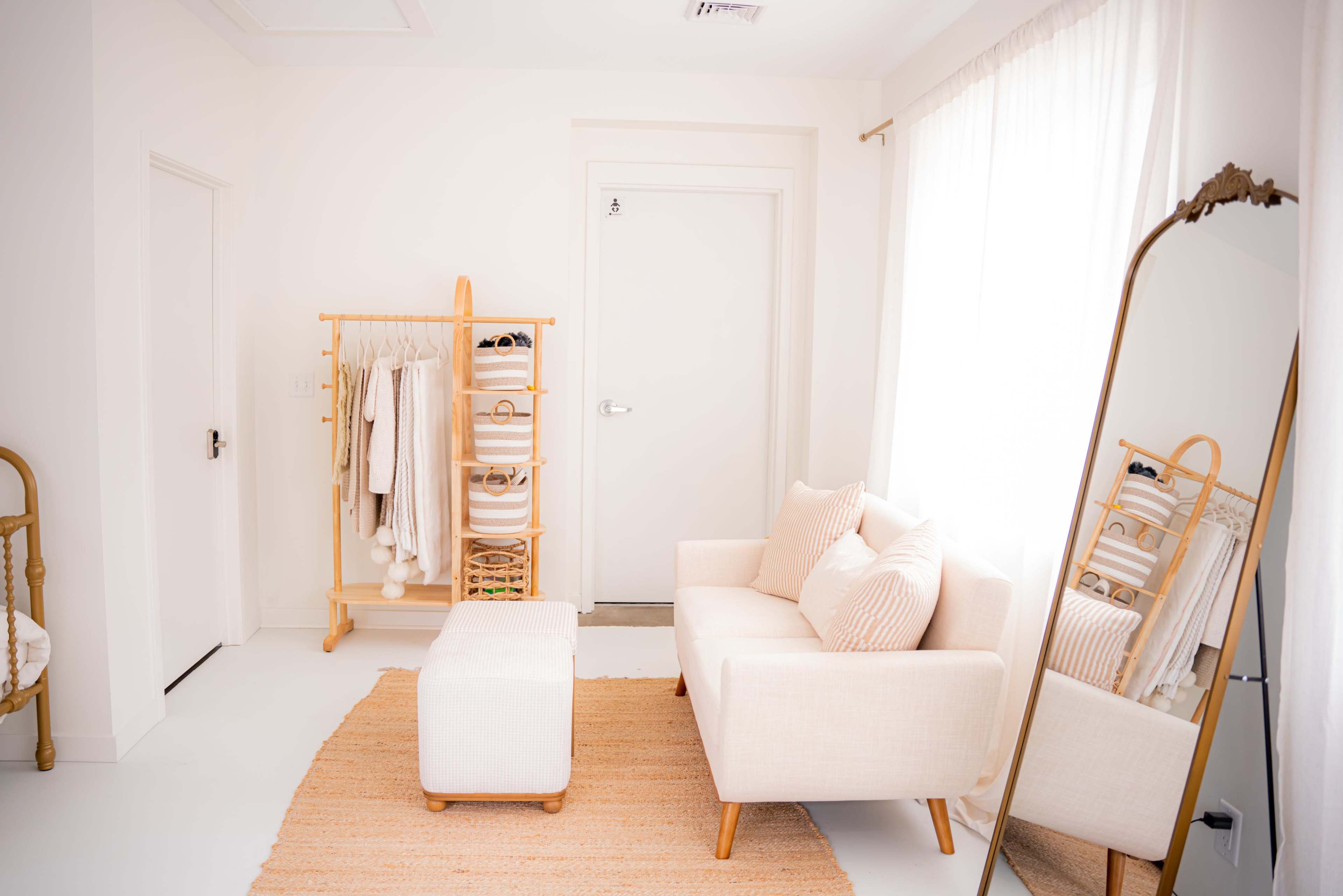 The image shows a light-colored room featuring a small sofa, a plush ottoman, a clothing rack with several items, and a large mirror.