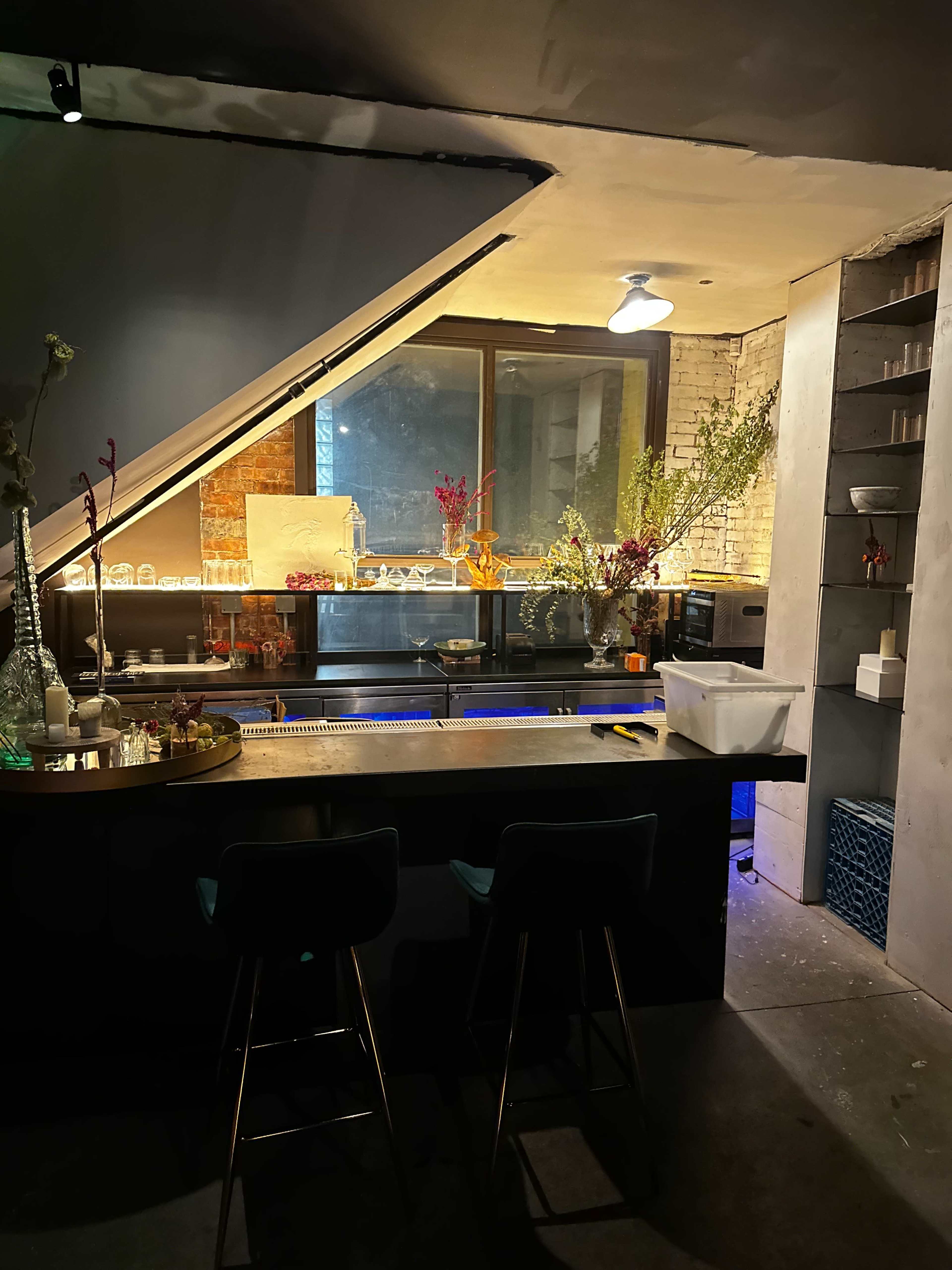 The image shows a dimly lit bar area with a sloped ceiling, a counter with two stools, and decorative flowers along the back wall.