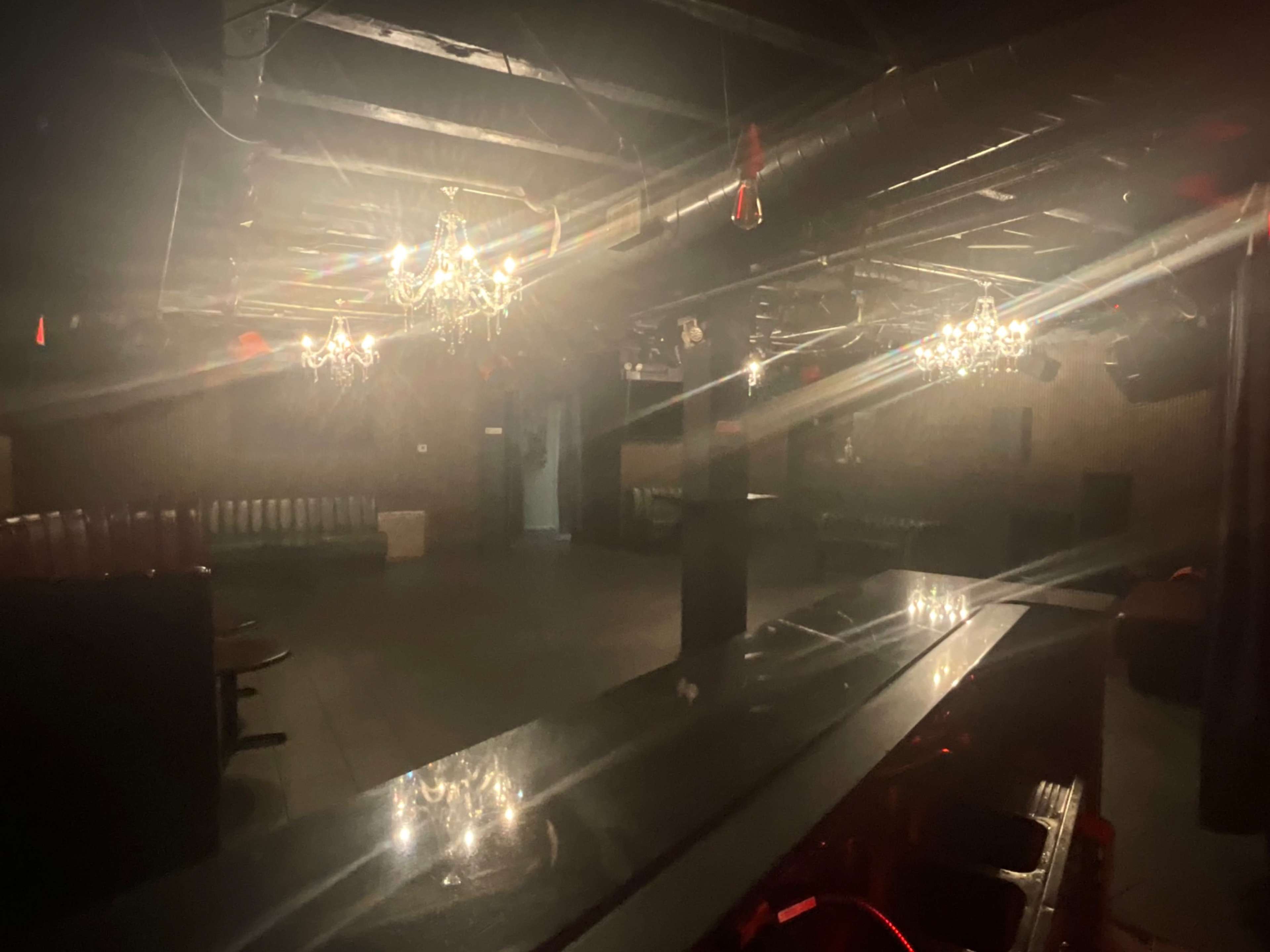 The image shows an empty bar area in a dimly lit venue with chandeliers and a polished counter.