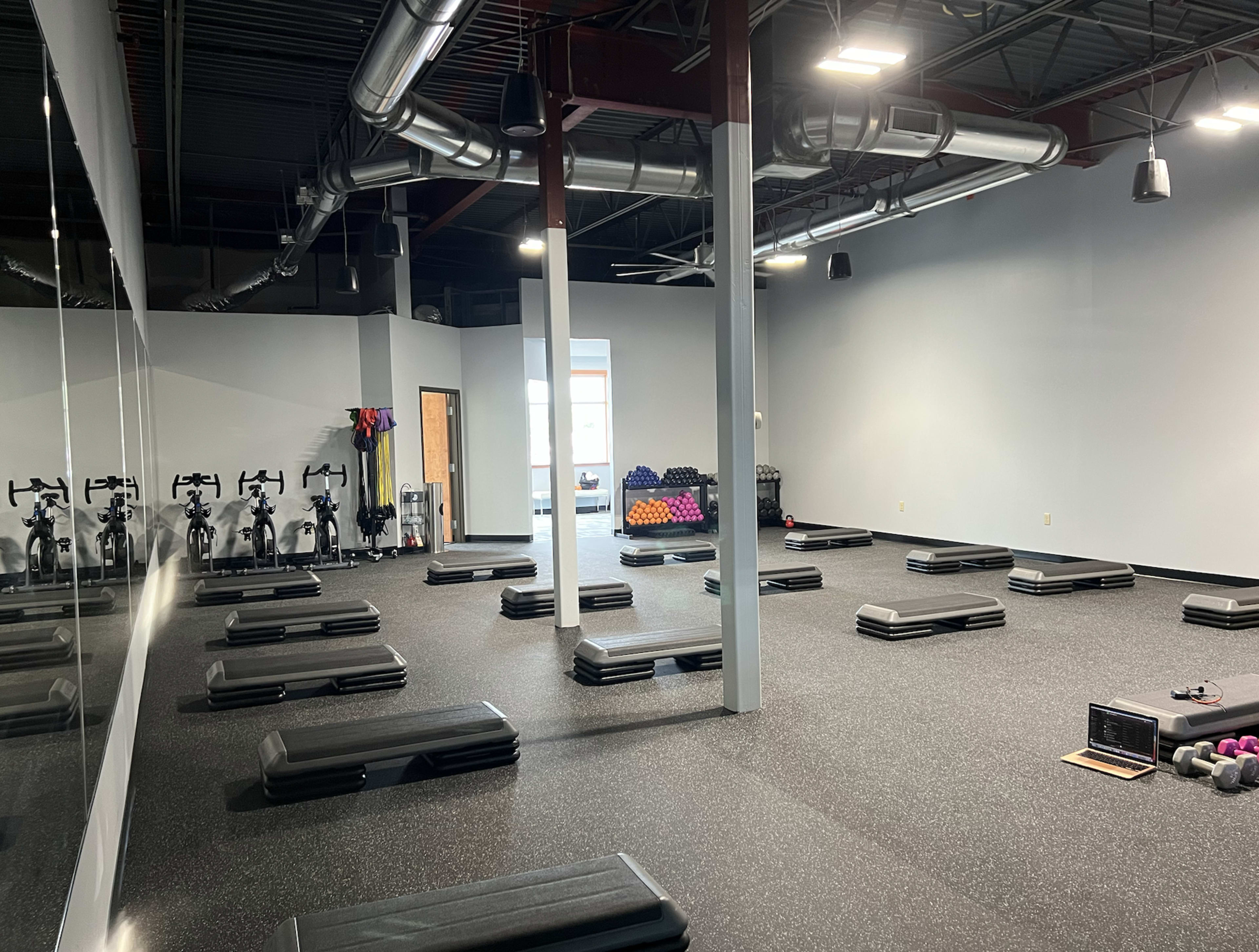 A spacious fitness studio with numerous step platforms arranged on the black rubber floor, exercise bikes lined up against the wall, and a few weights scattered in one corner.