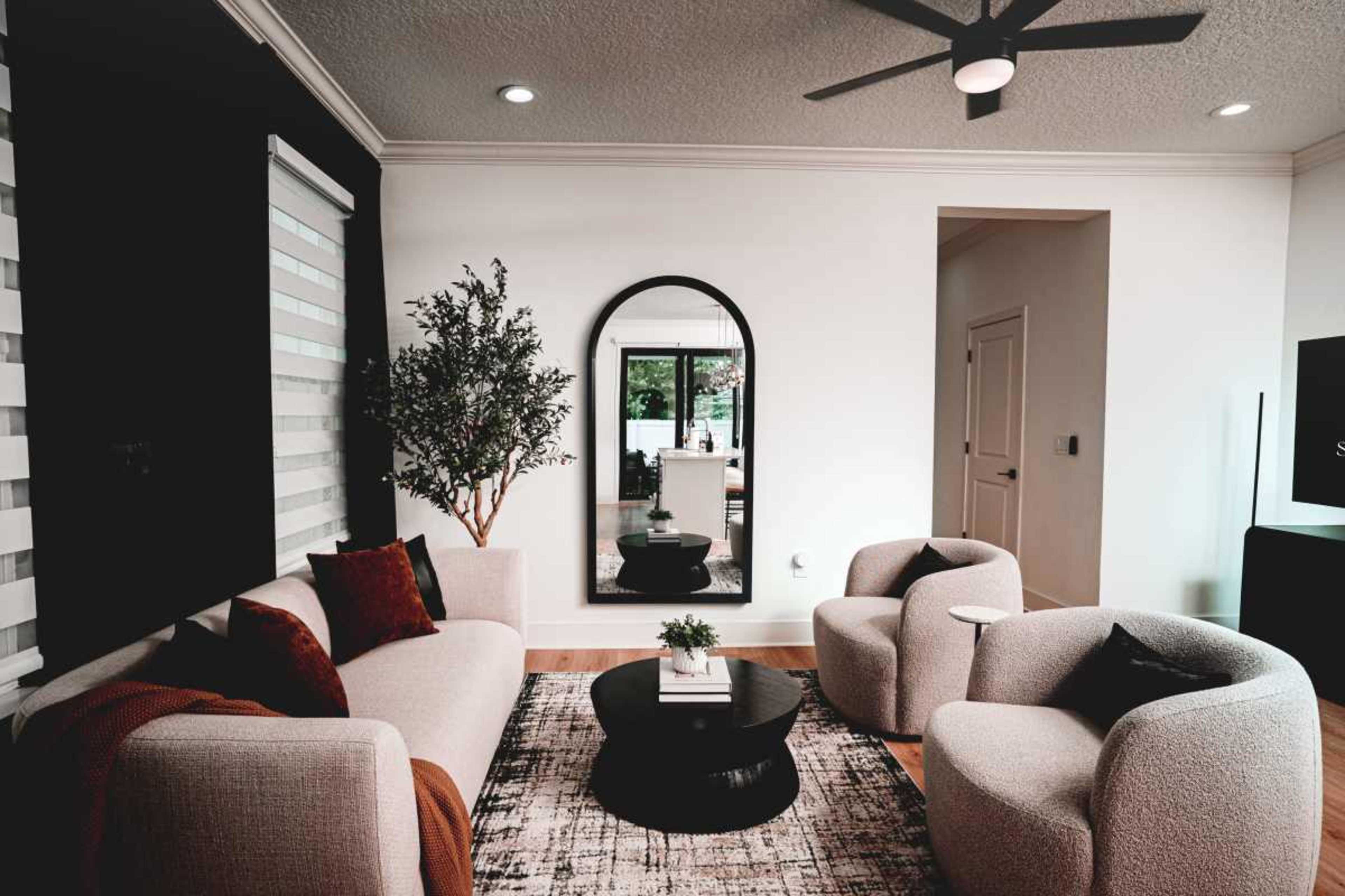 The image shows a modern living room with a light-colored sofa, two rounded armchairs, a circular coffee table, and a large mirror on the wall opposite a window.