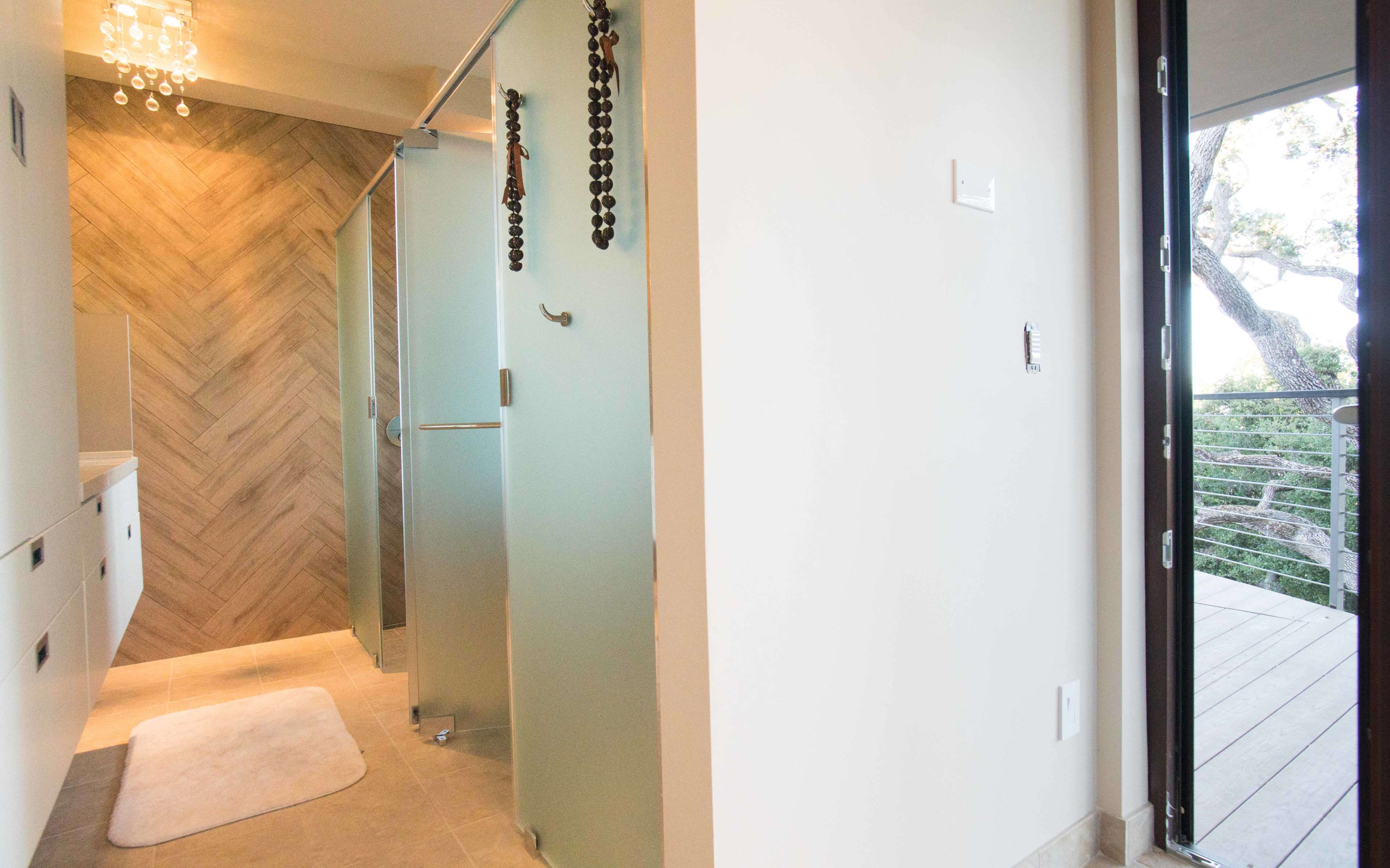 The image shows a modern bathroom on the left with a glass shower and herringbone tile, and a blank wall on the right leading to a balcony with a railing.