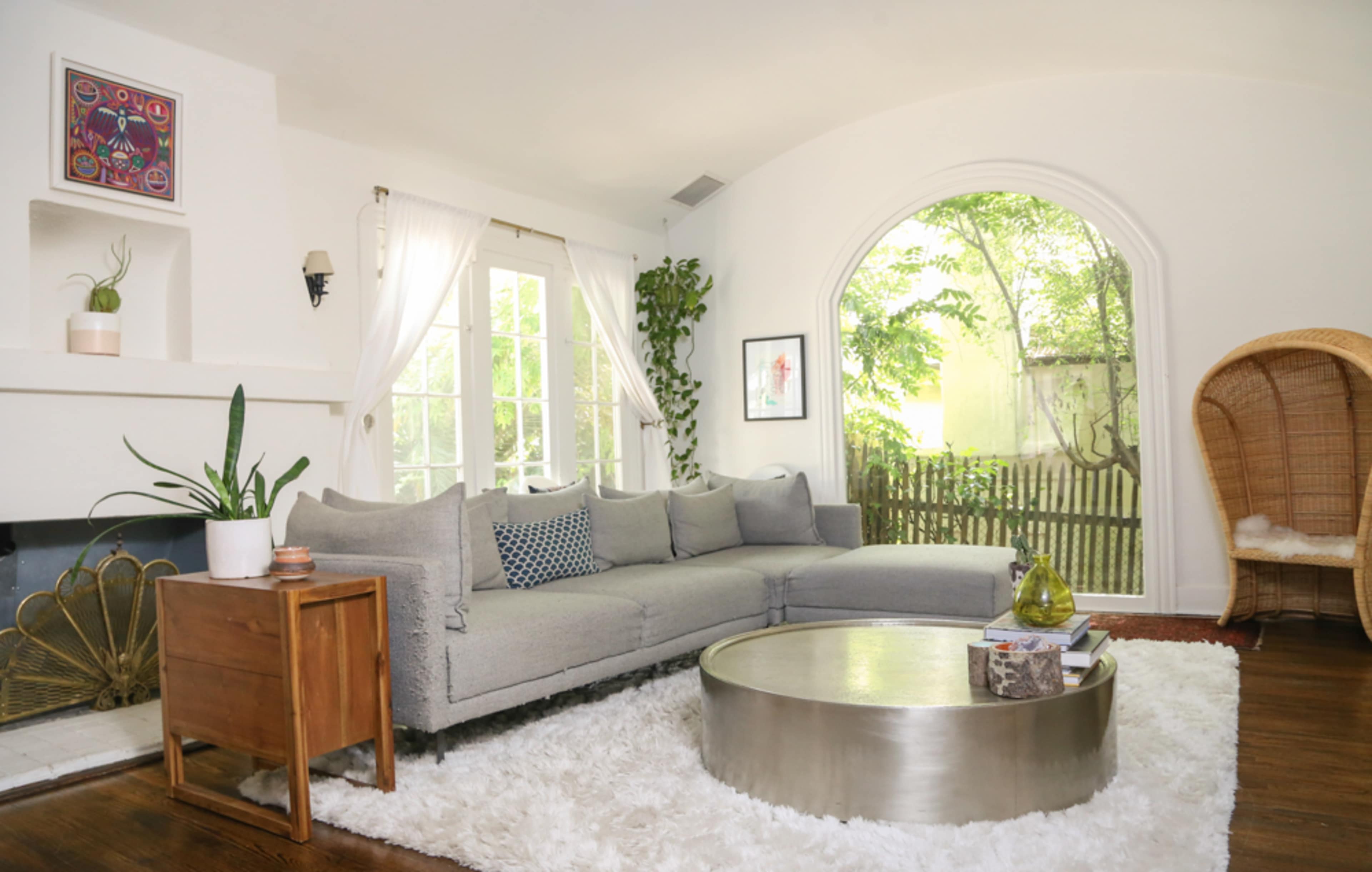 The image shows a cozy living room with a gray sectional sofa, a round coffee table, and a large window overlooking a green outdoor space.