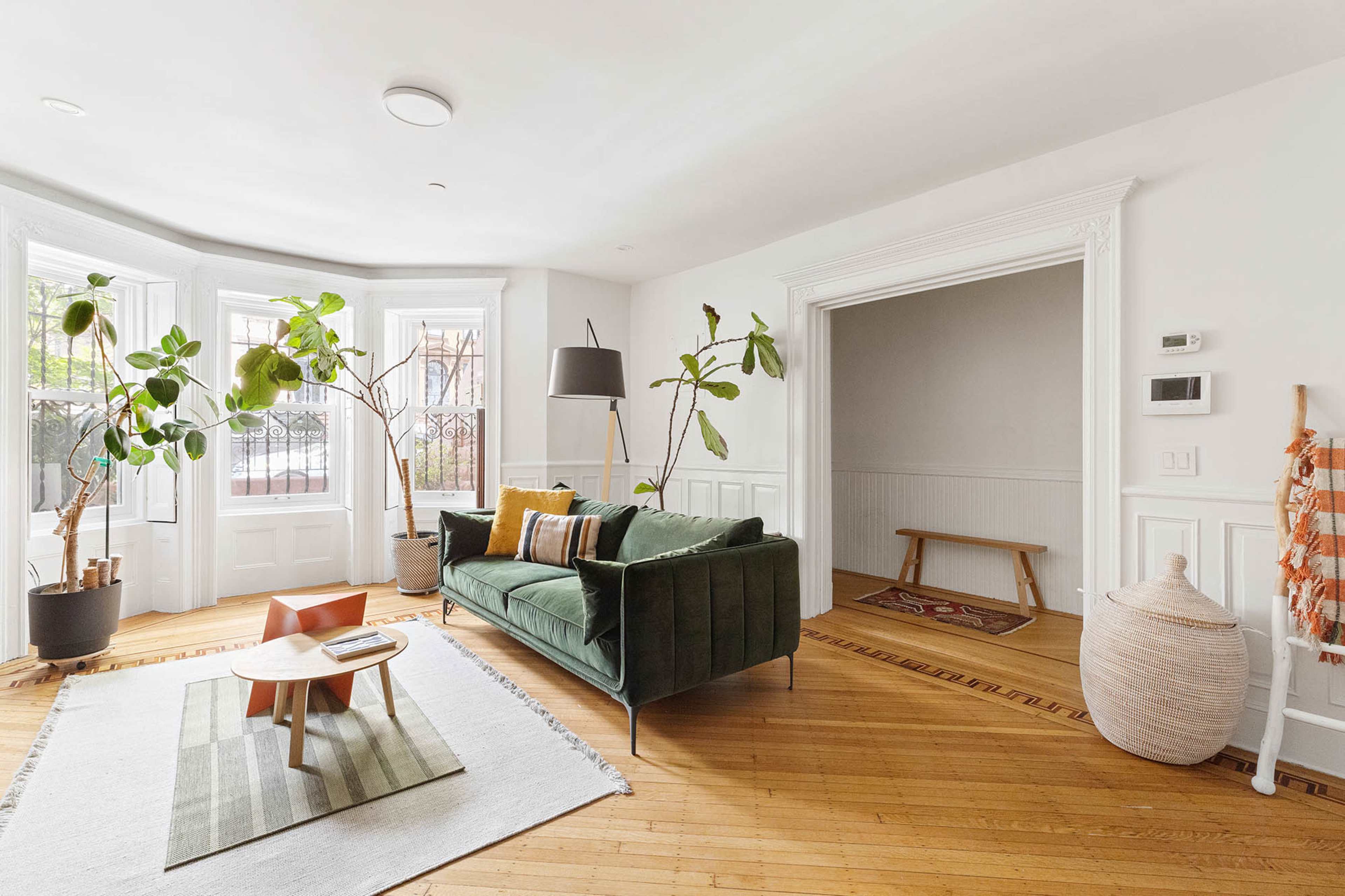 The image shows a bright living room with a green couch, a coffee table on a rug, and large plants near bay windows.