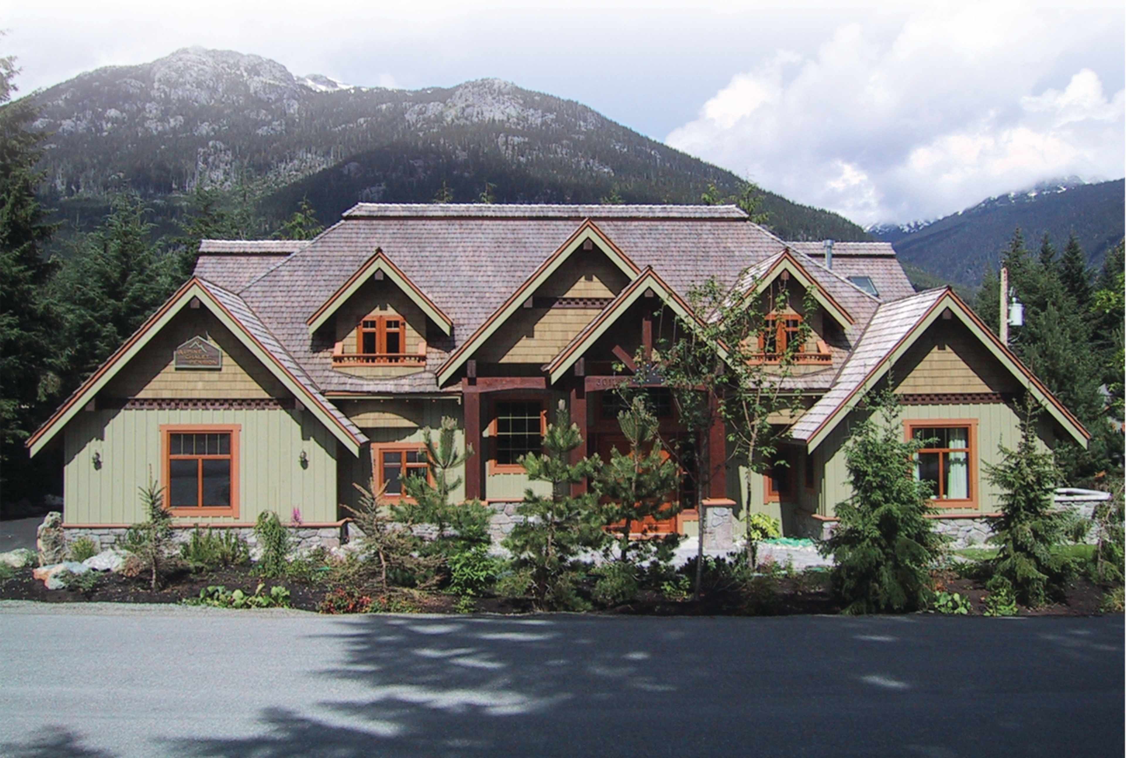 A large, multi-roofed house featuring a combination of green and tan siding, set against a backdrop of mountains and trees.