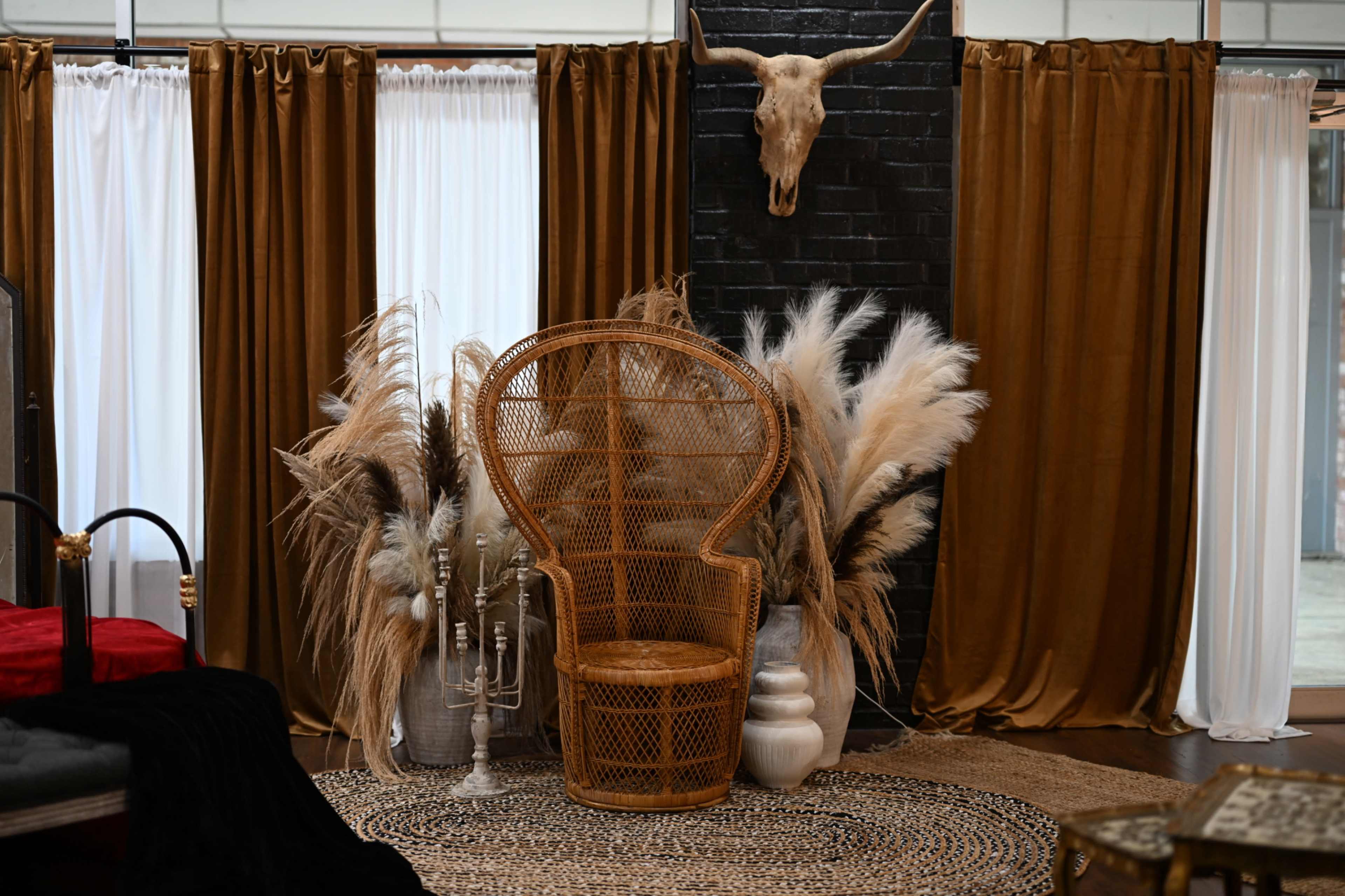A rattan chair surrounded by dried pampas grass and a decorative skull mounted on the wall, all framed by dark and light curtains.