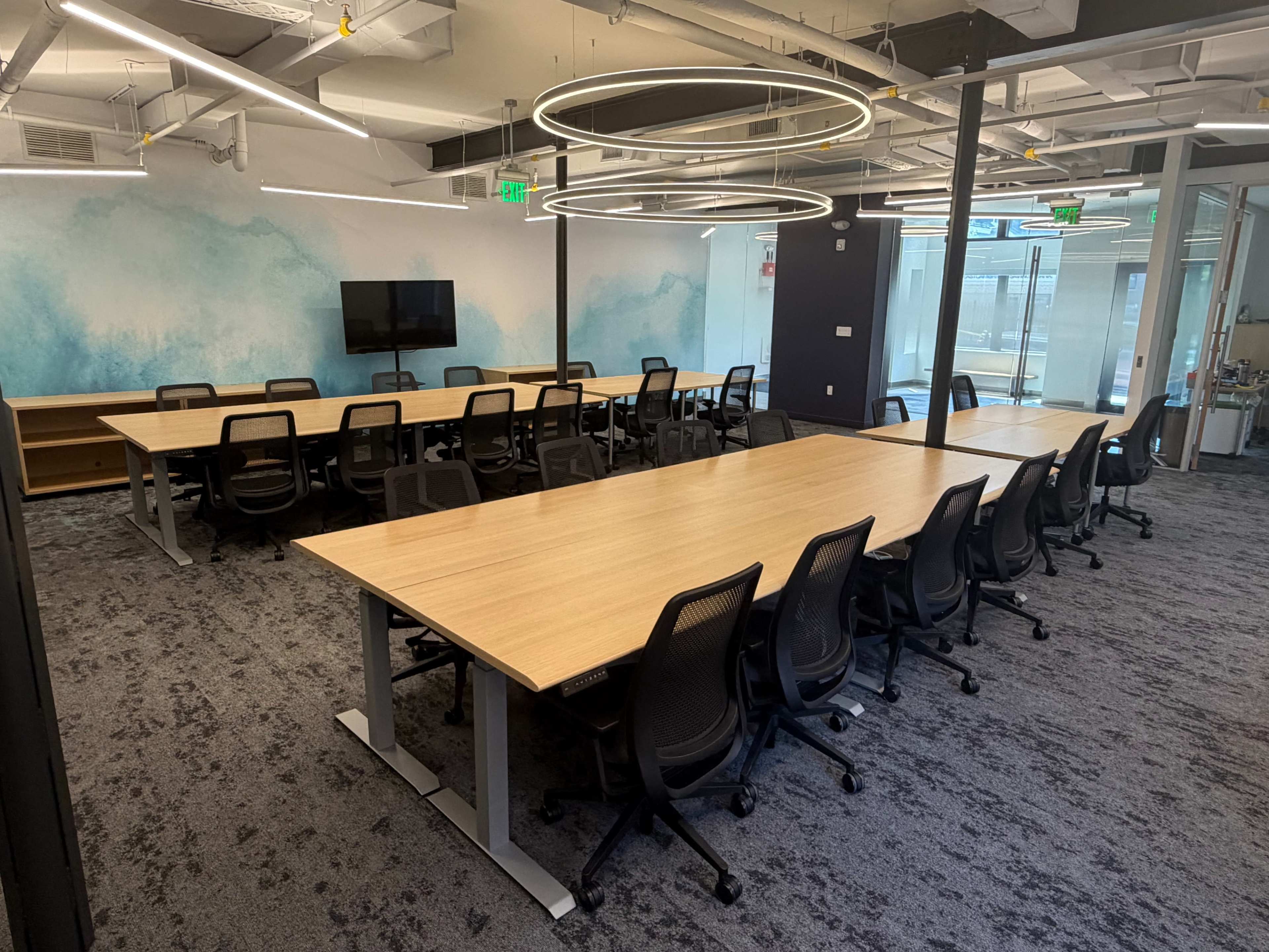 The image shows a modern conference room with several long wooden tables arranged in a U-shape, surrounded by black swivel chairs and large windows.