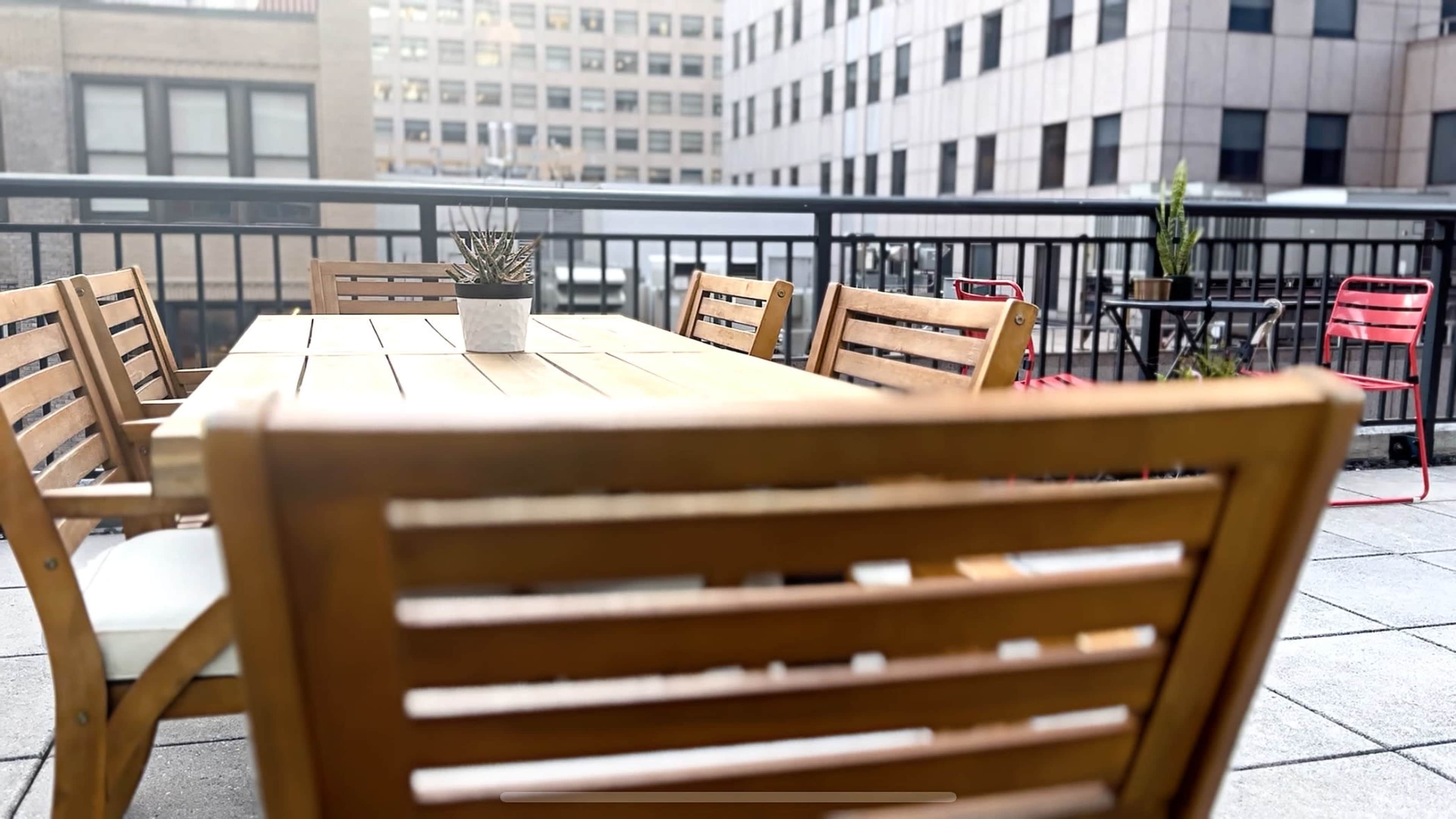 A wooden dining table is surrounded by several chairs on a rooftop terrace with a view of nearby buildings.