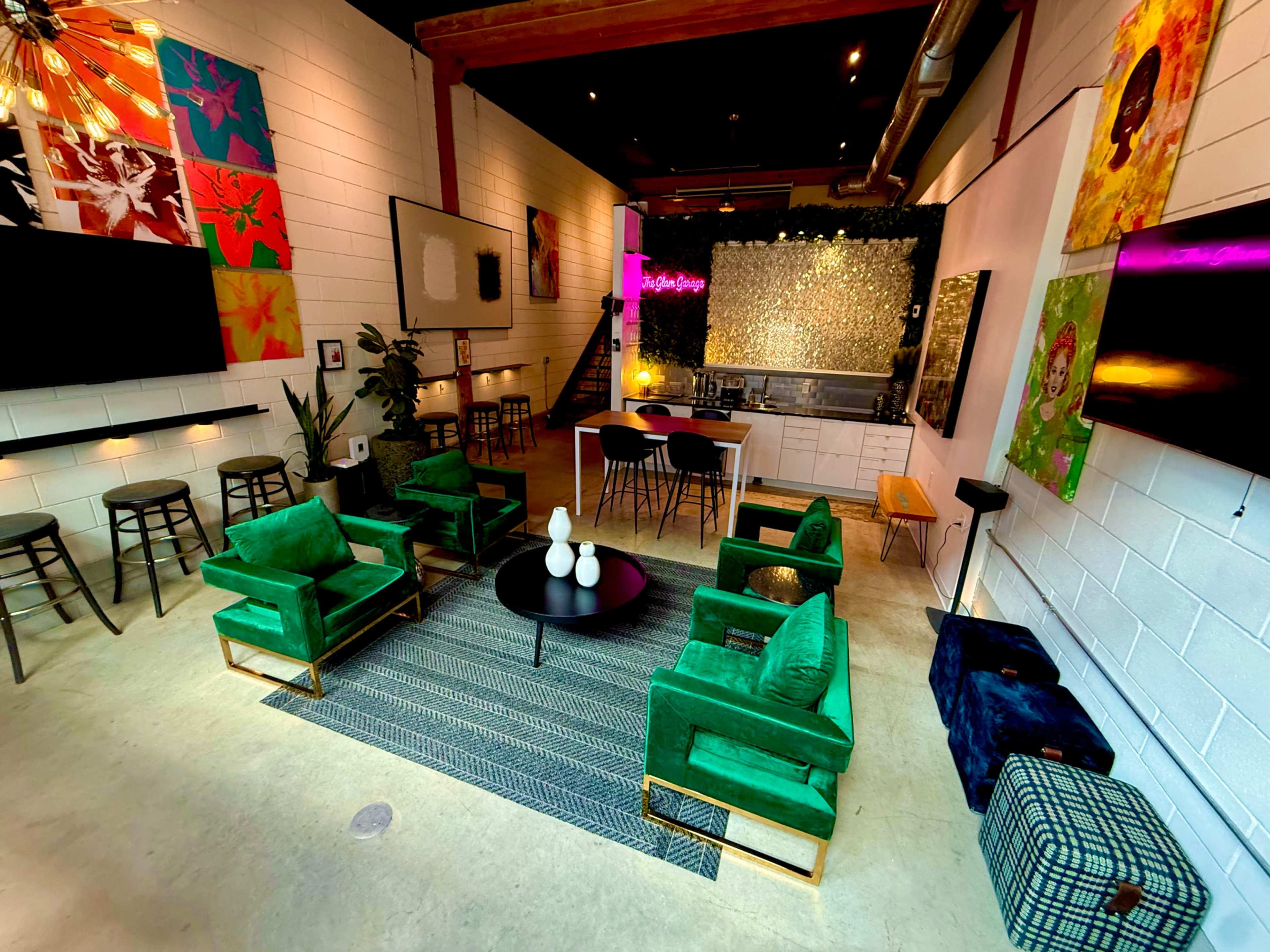 The image shows a modern, open-concept lounge area featuring green armchairs, a round black coffee table, and a kitchen visible in the background, adorned with colorful artwork on the walls.