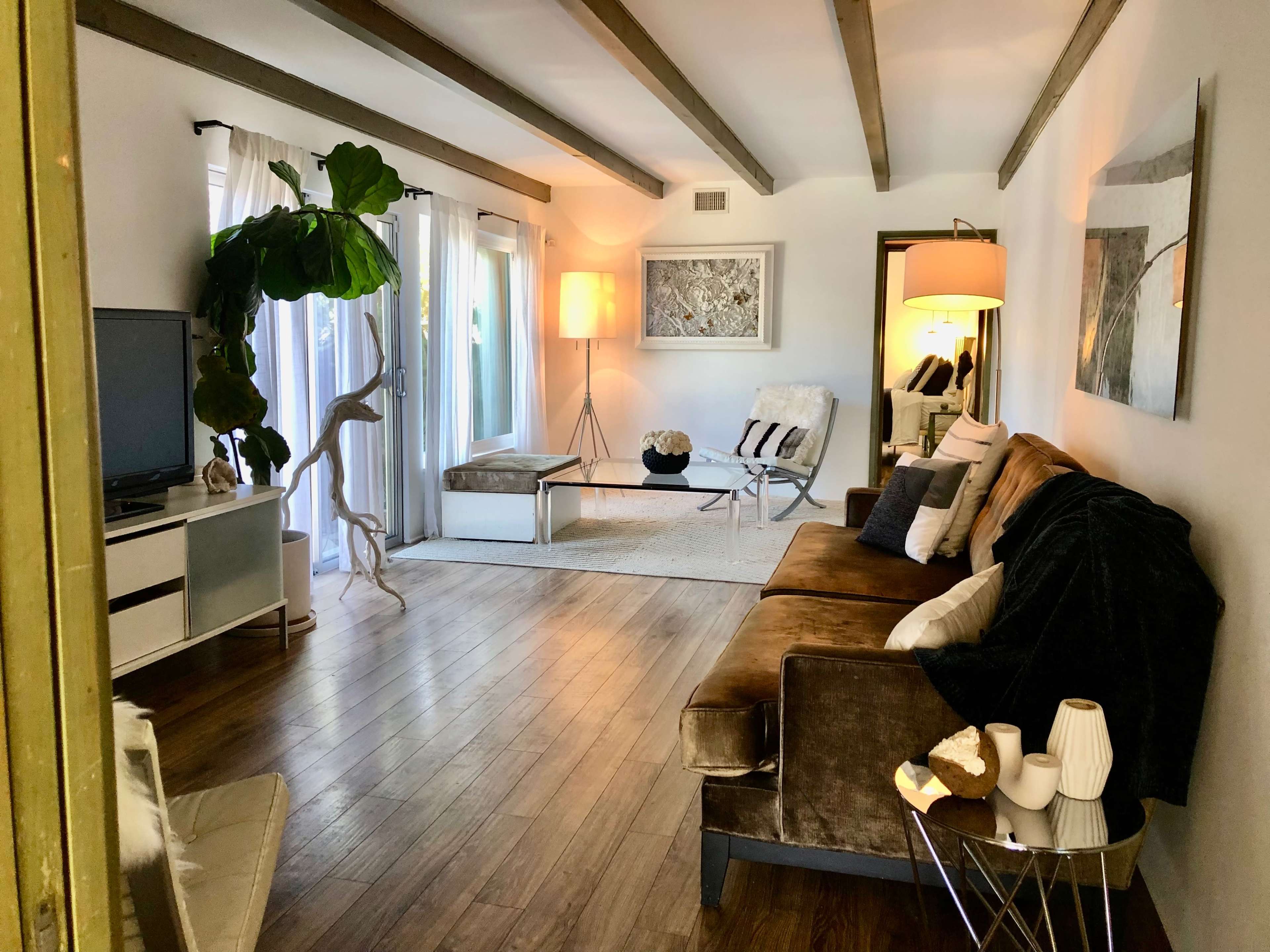 A living room with a sofa, coffee table, floor lamp, and large windows, featuring wooden beams on the ceiling and a view into a mirrored space.