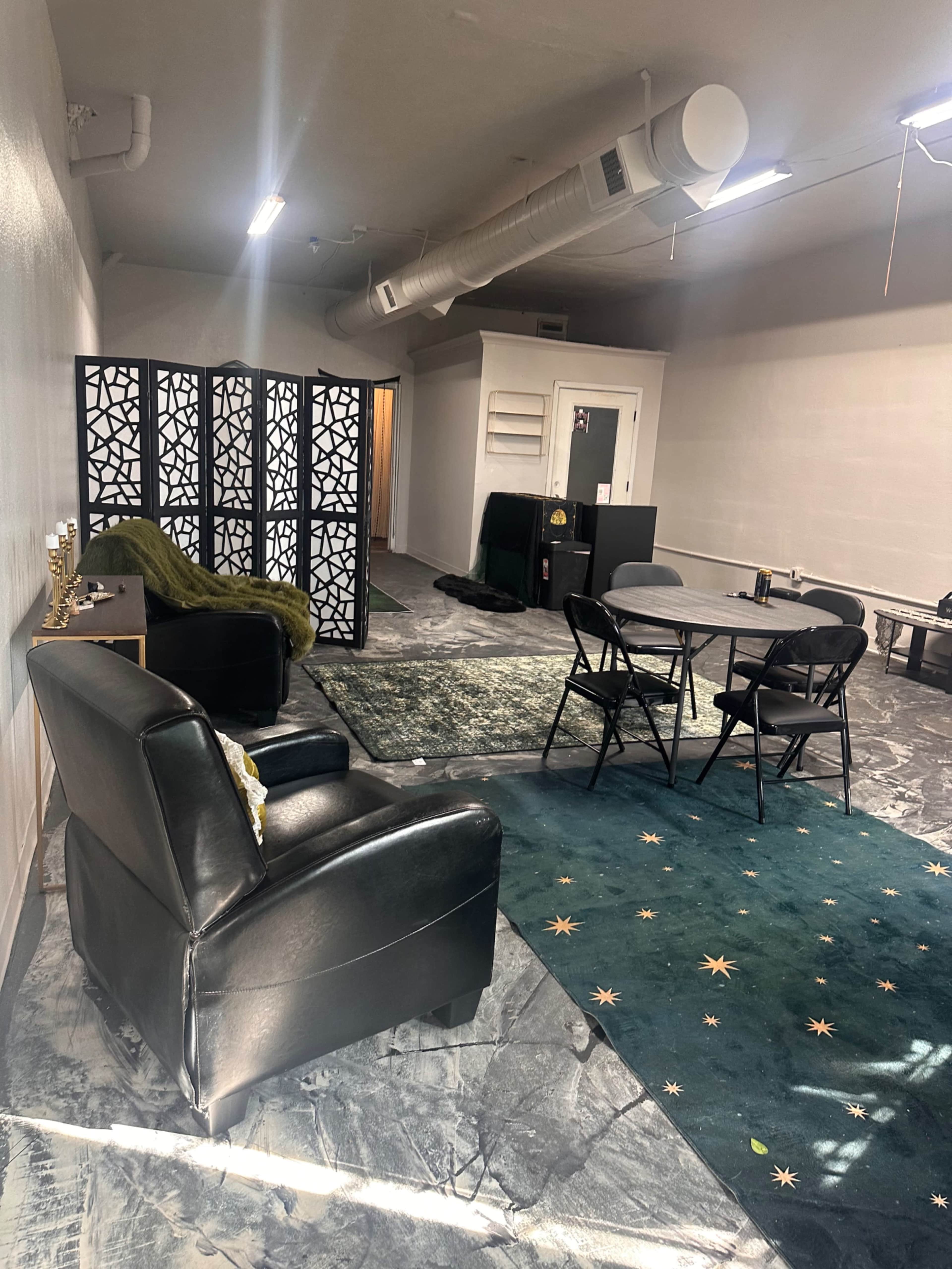 The image shows a spacious room with a seating area featuring black chairs, a patterned folding screen, and a circular table surrounded by black folding chairs, all set on a carpeted floor with decorative design elements.