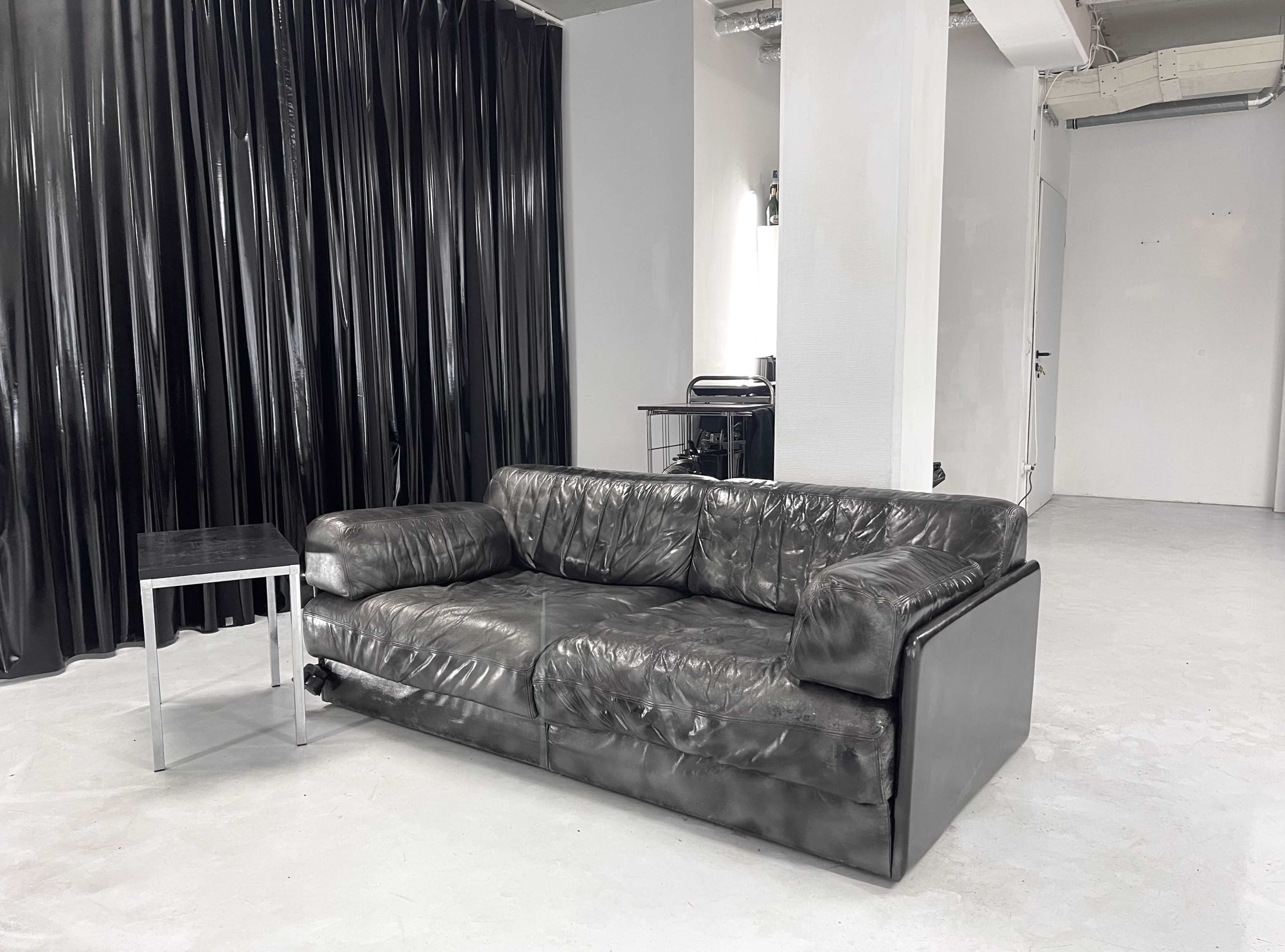 A dark leather sofa is placed next to a small metallic side table in a minimalist room with a black curtain backdrop.