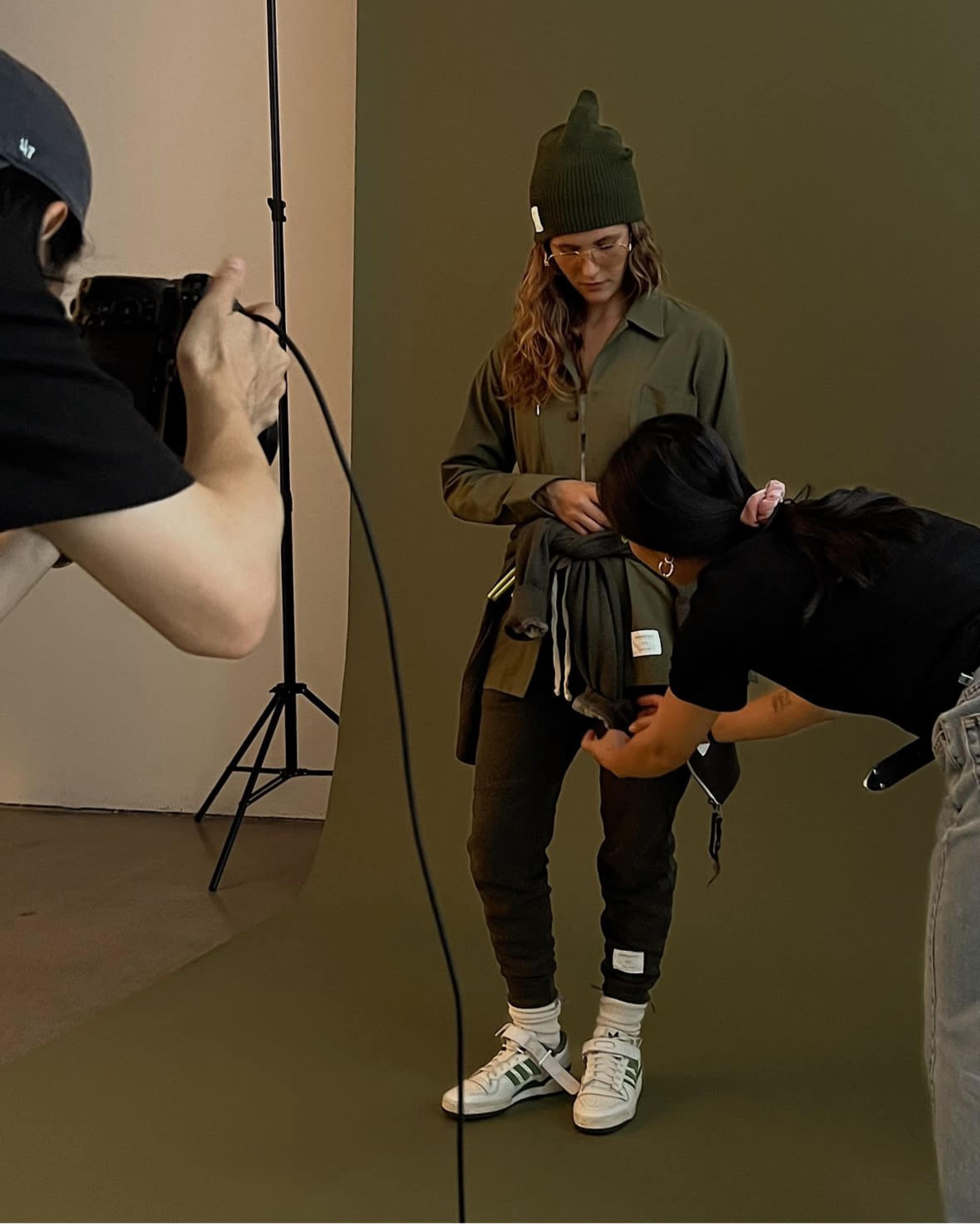 A model poses in a green outfit while a stylist adjusts her clothing in a studio setting with a neutral backdrop.