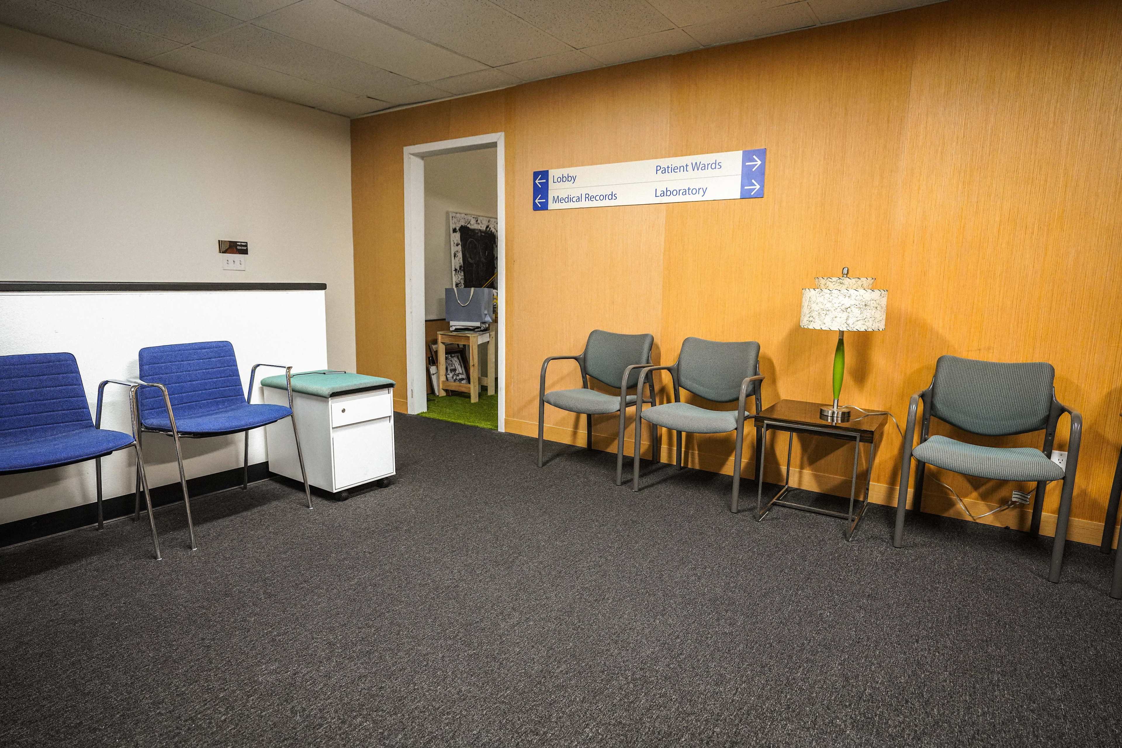 The image shows a waiting area with several chairs arranged around a small table, a trash bin, and directional signs on the wall.