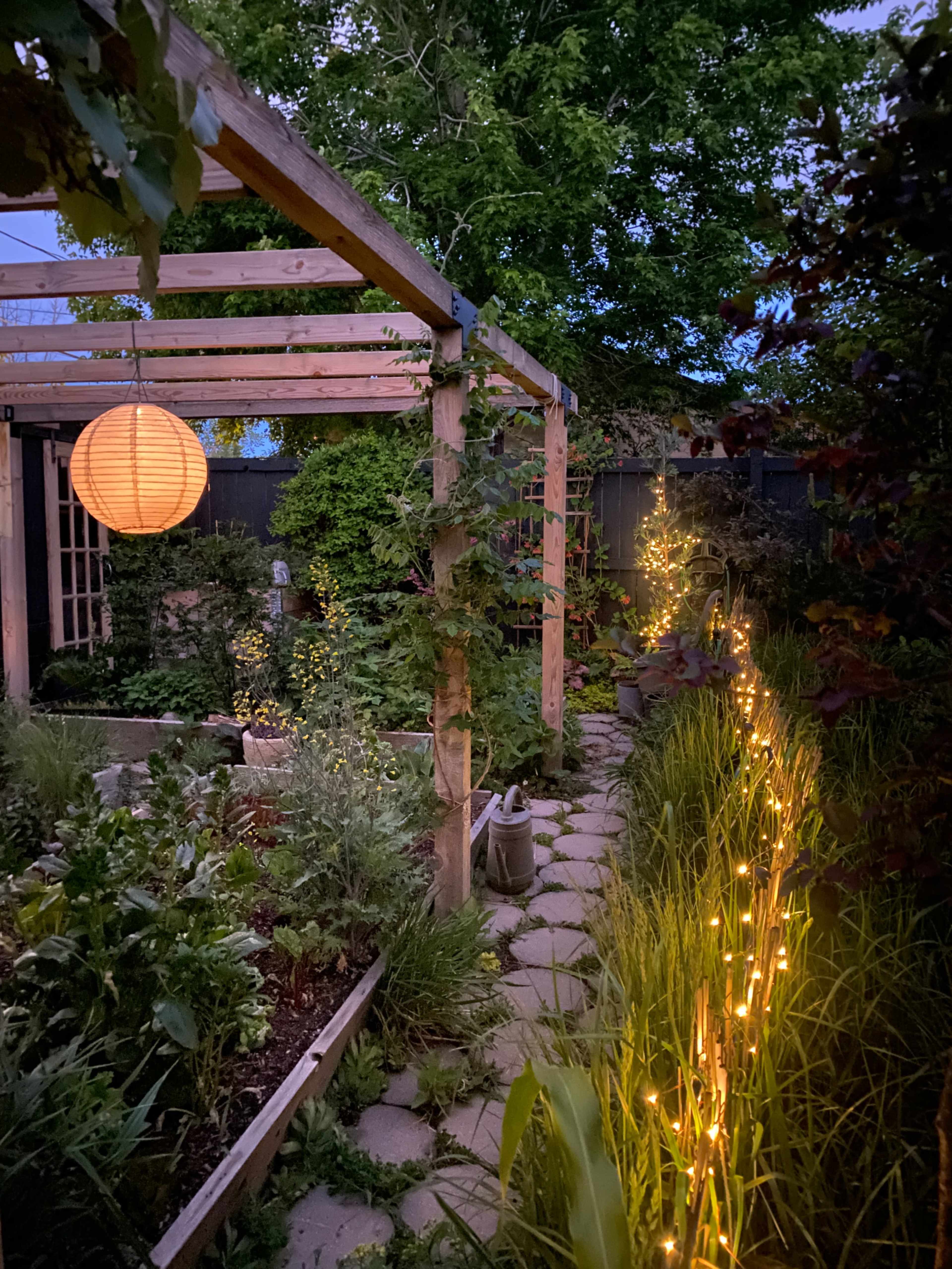 A well-lit garden path lined with glowing lights leads under a wooden arbor with a lantern hanging nearby.