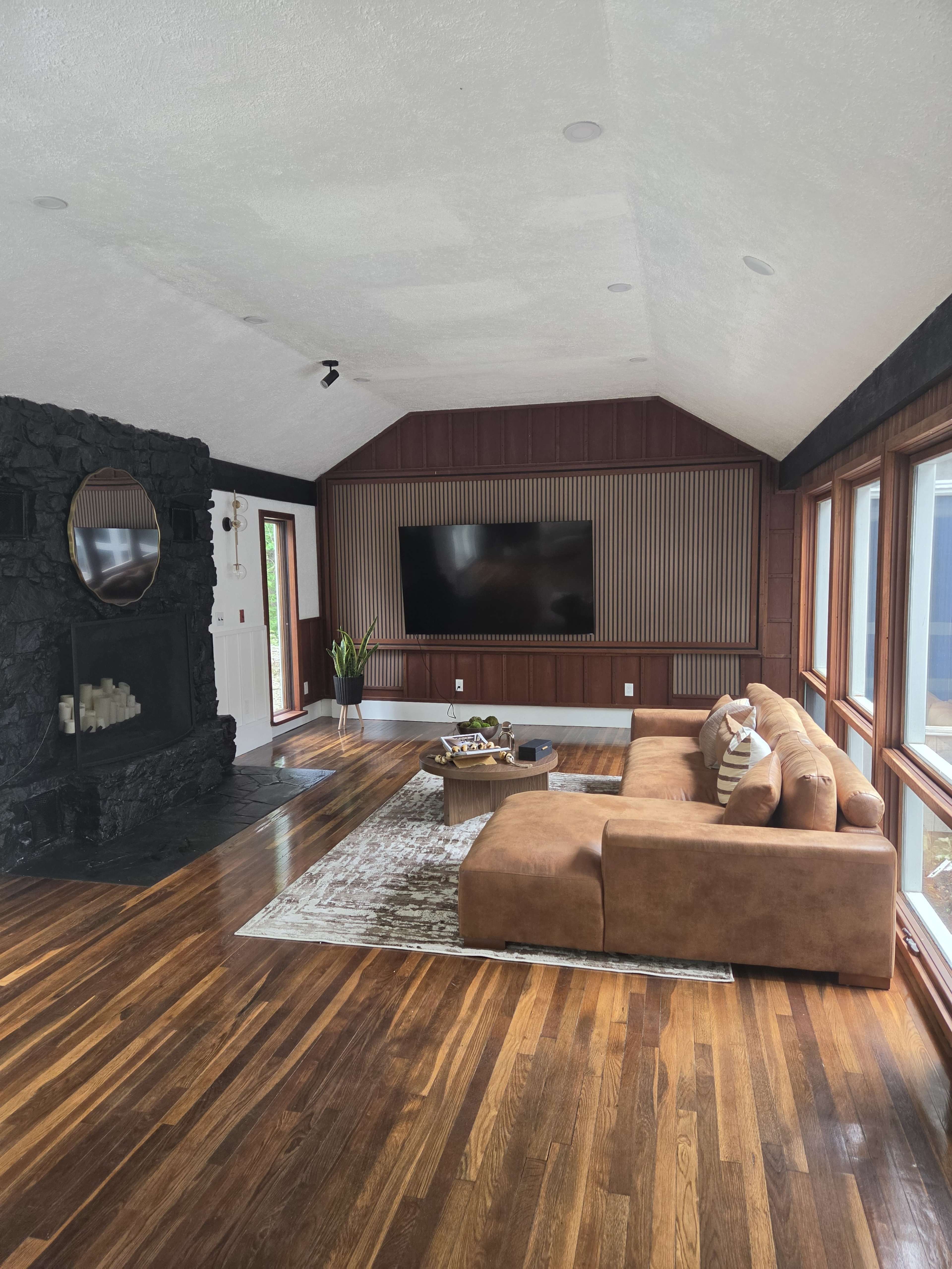 The image shows a spacious living room with a large brown sectional sofa, a coffee table, and a stone fireplace, featuring large windows that allow natural light to enter the space.