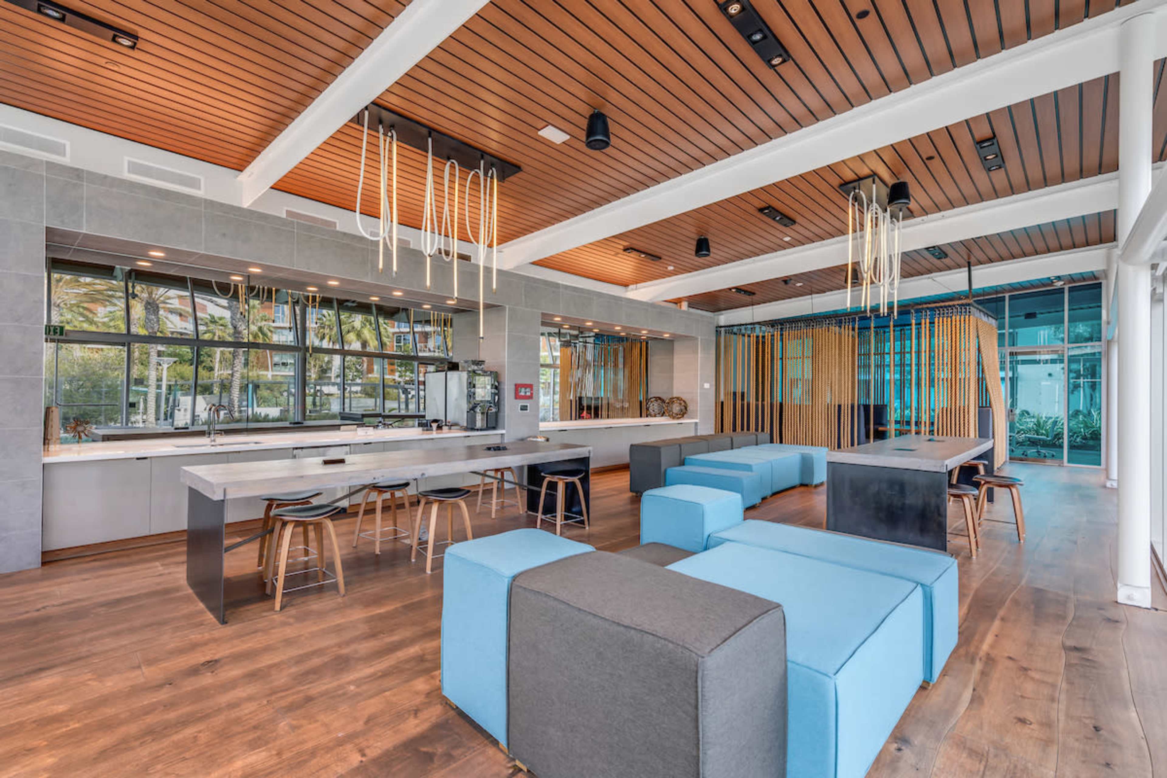 A modern interior space with wooden floors, large windows, and a combination of blue and gray seating options arranged around a central island and tables.