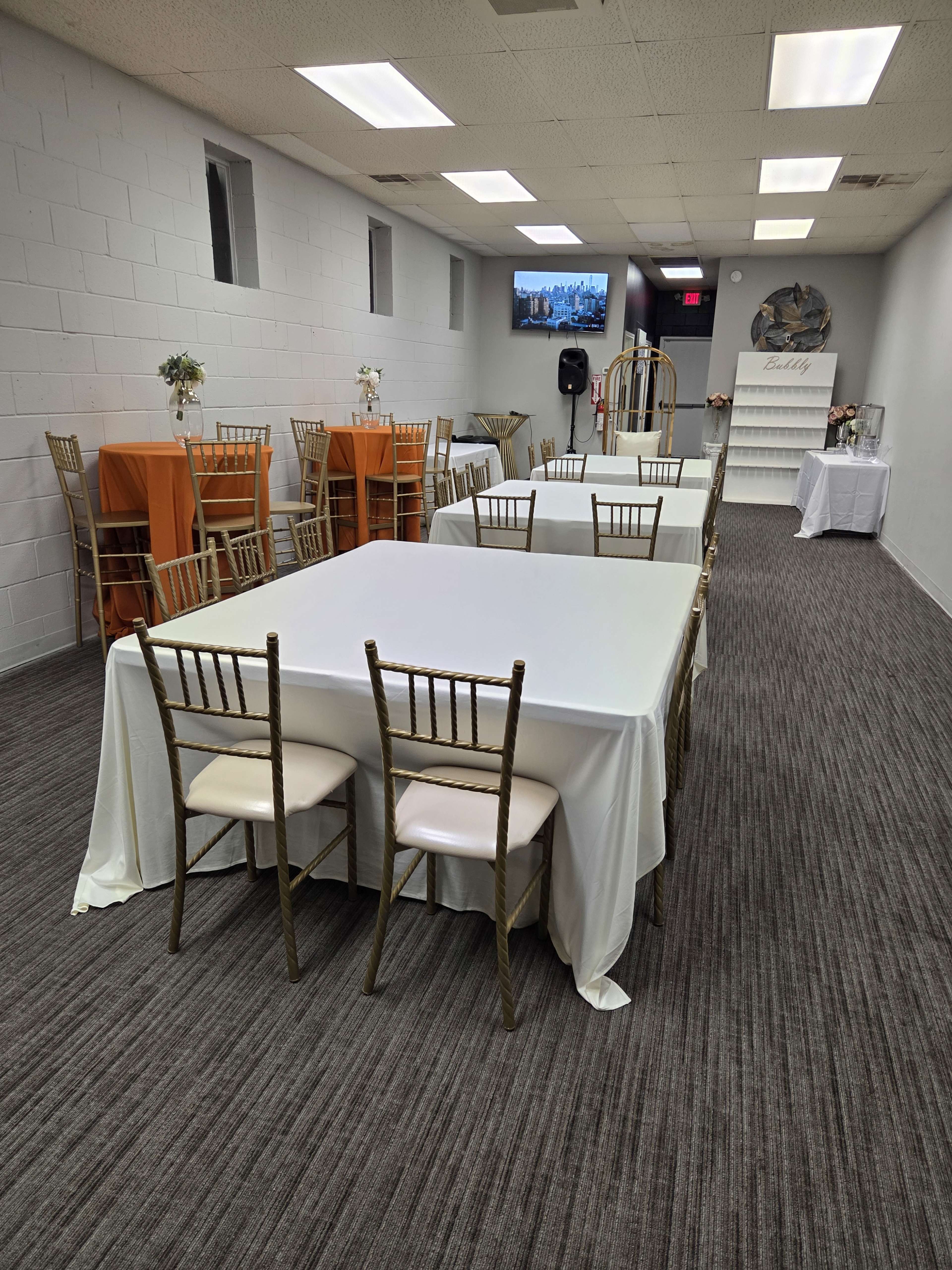 The image shows a spacious event room with white and orange tablecloths on various tables, gold chiavari chairs, and a small stage area in the background.