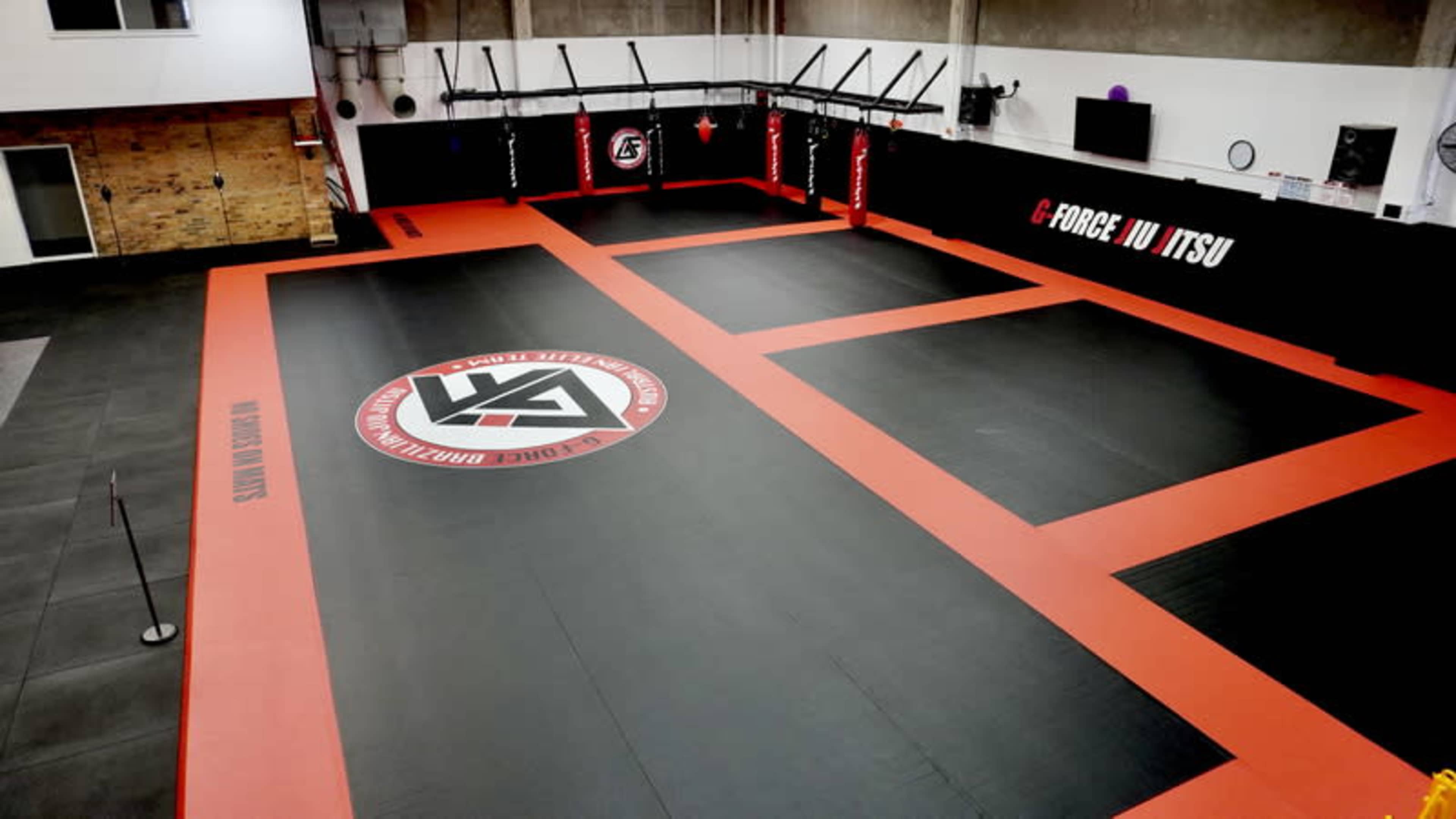 The image shows a martial arts training facility with a large black and red mat area, featuring marked training zones and a logo in the center.