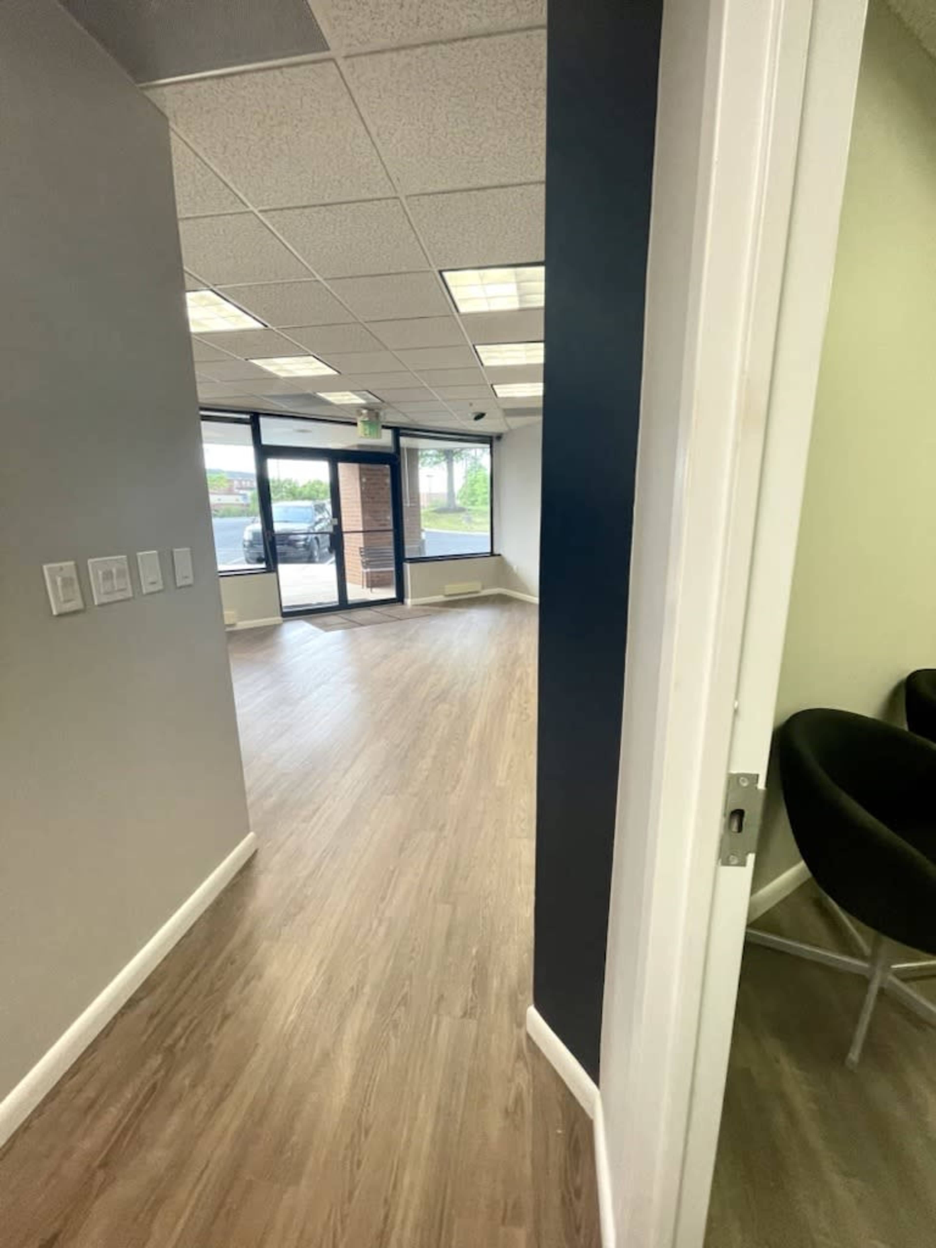 A door opens to a spacious, well-lit room with wooden flooring and large windows overlooking an outdoor area.