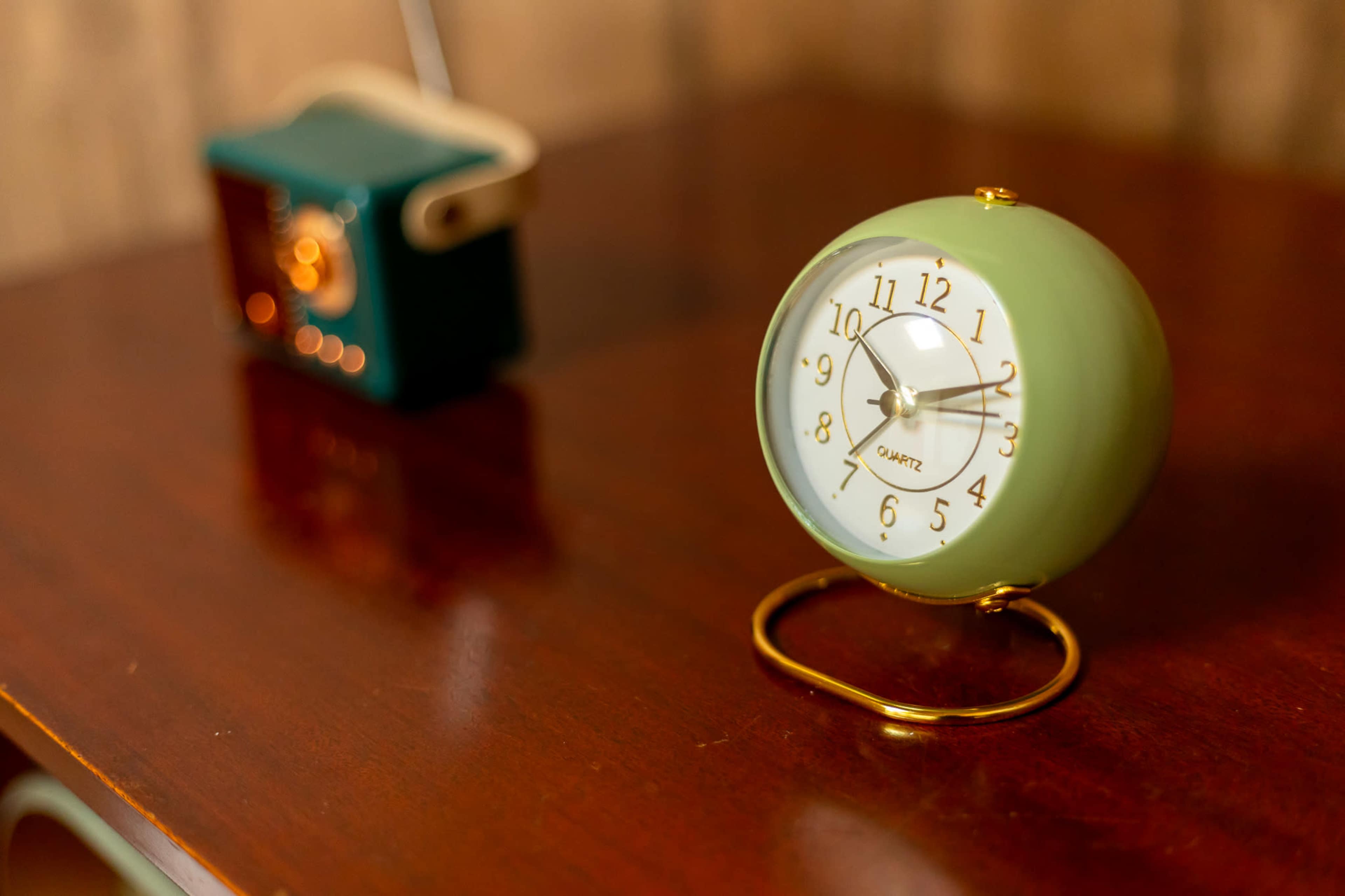 A round, mint green alarm clock with a white face and gold accents sits on a wooden surface next to a small retro-style radio.
